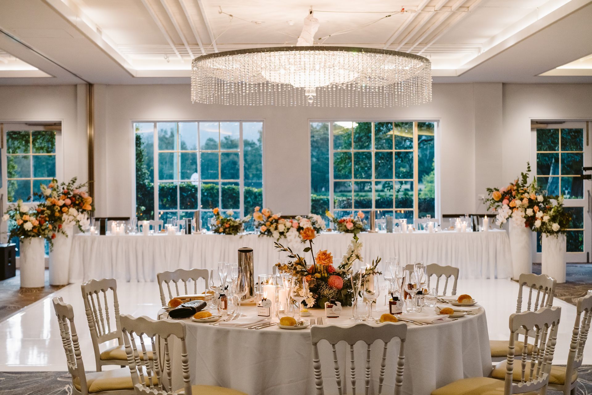 Elegant wedding reception with round table, flowers, chandelier, and outdoor view through windows.