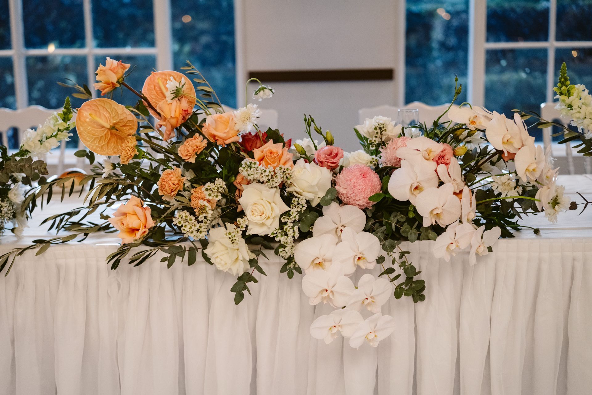 Floral arrangement on a white draped table with white chairs, next to windows.
