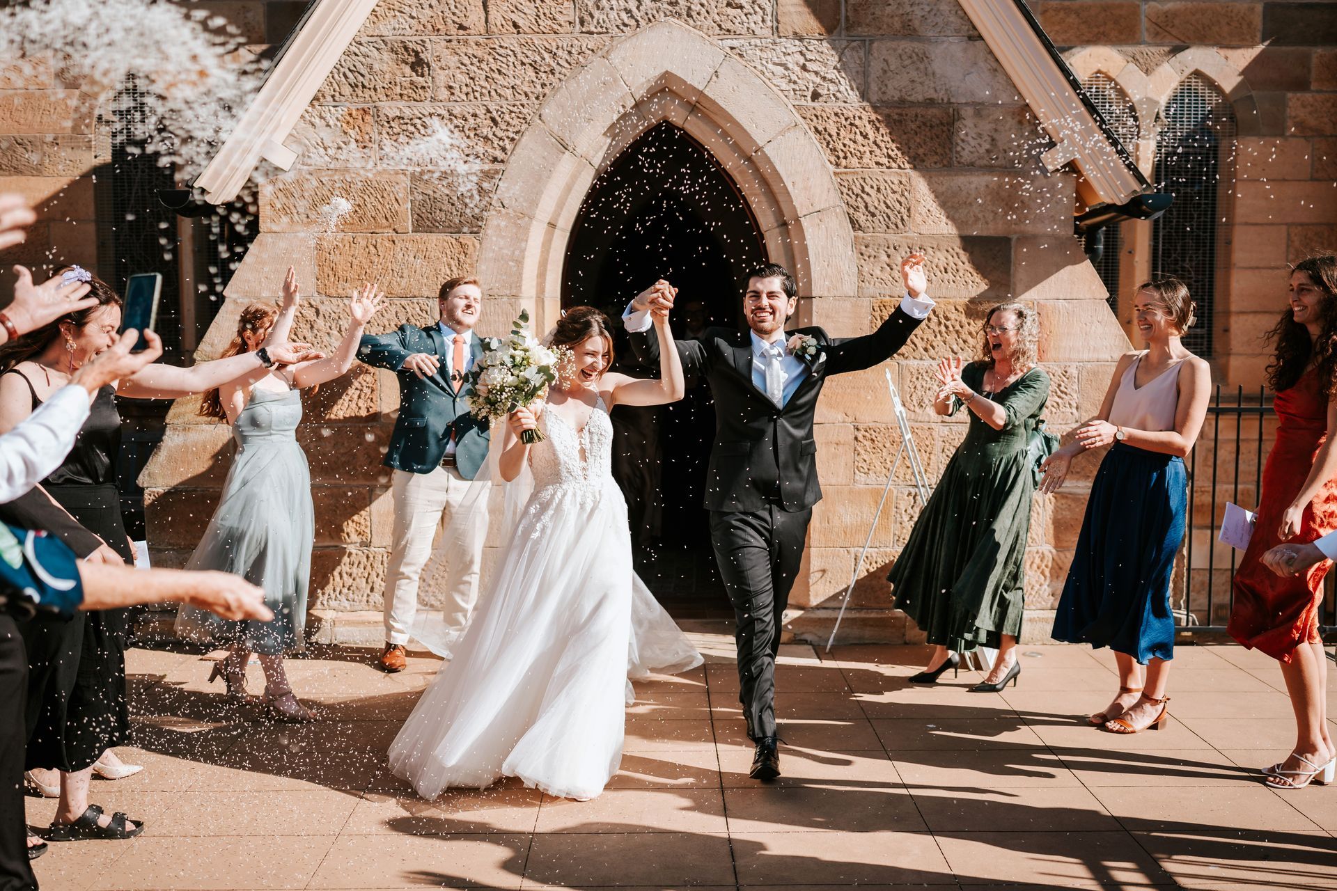 Newlyweds exit a stone church, showered with confetti by cheering guests.
