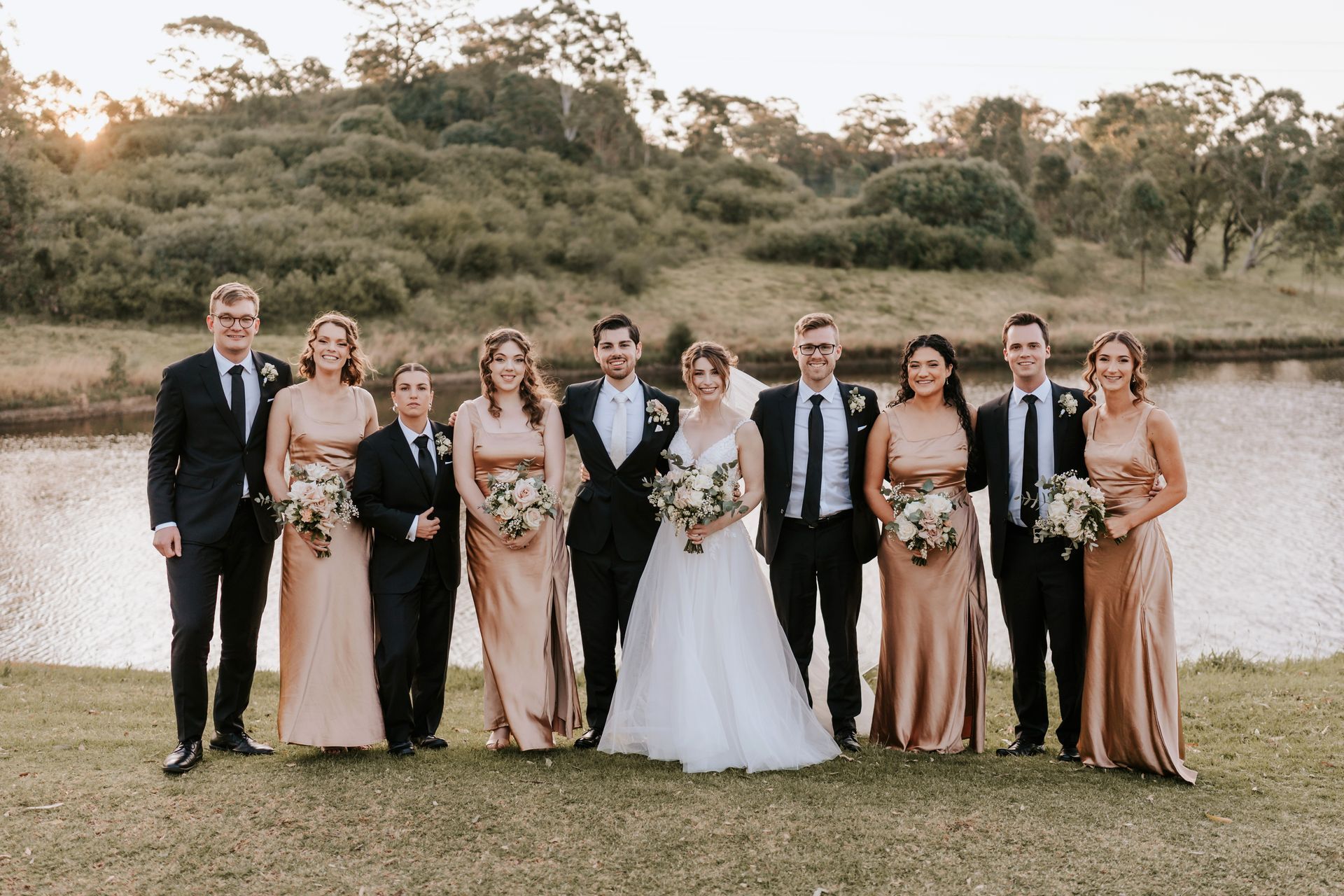 Wedding party poses by a lake; bride and groom centered, attendants in gold dresses/black suits.
