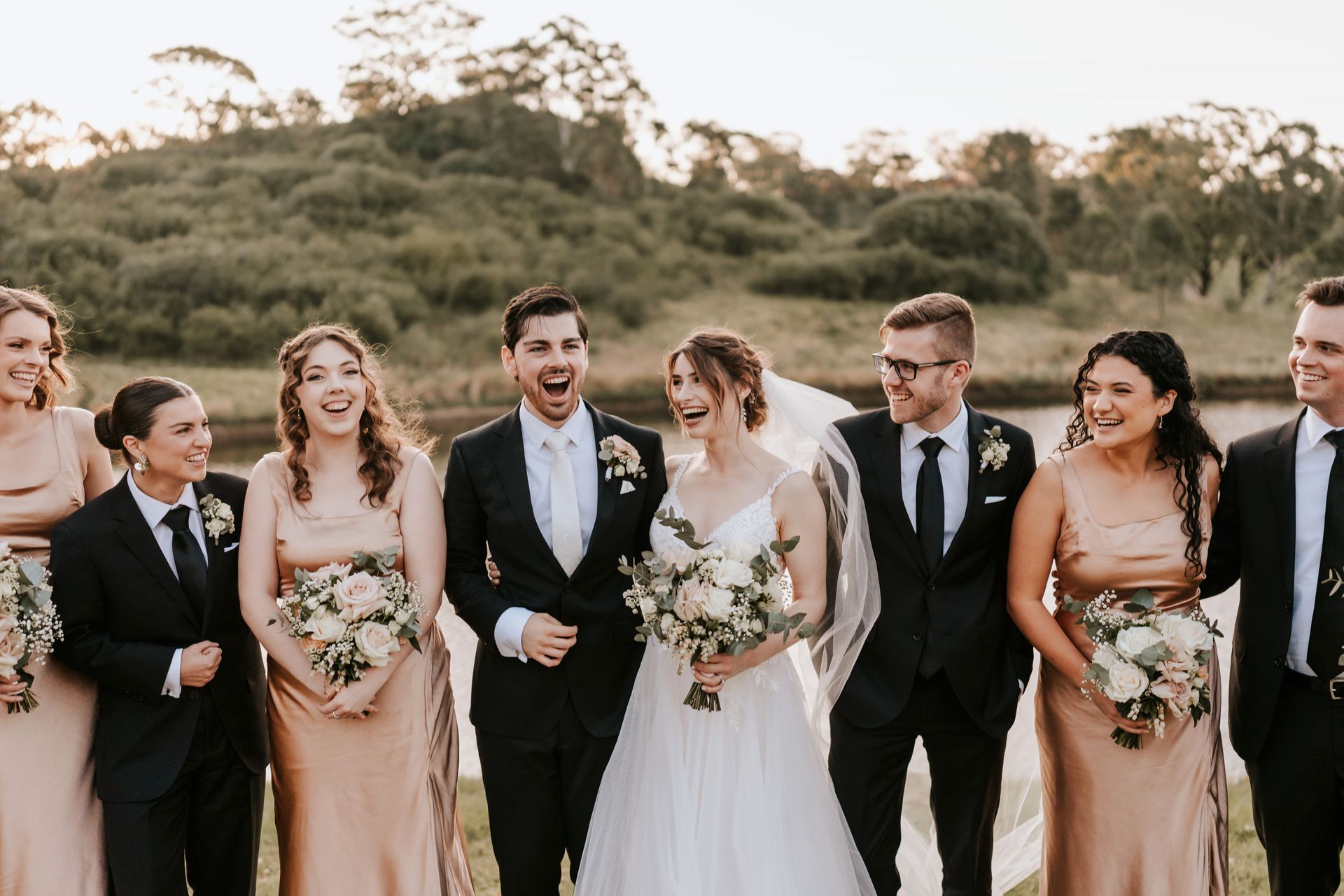 Wedding party: Bride and groom with bridesmaids and groomsmen, smiling by water.