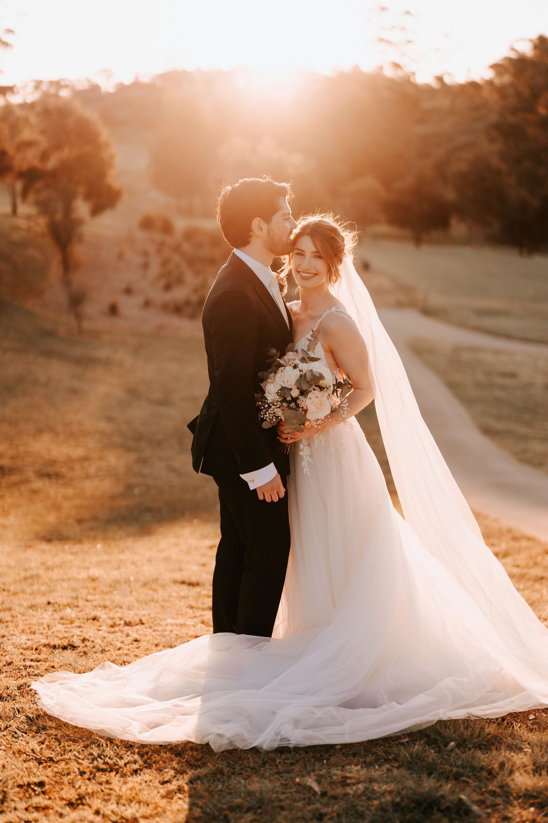 Bride and groom embracing in a field at sunset. The bride wears a white dress and veil, the groom in a black suit.