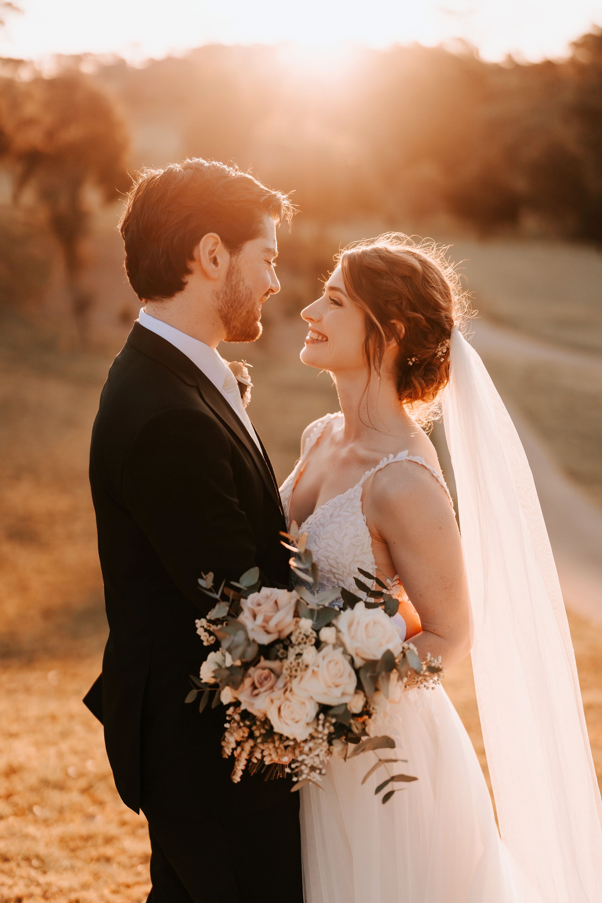 Bride and groom smiling at each other, outdoors. Bride holds bouquet, sunset in background.