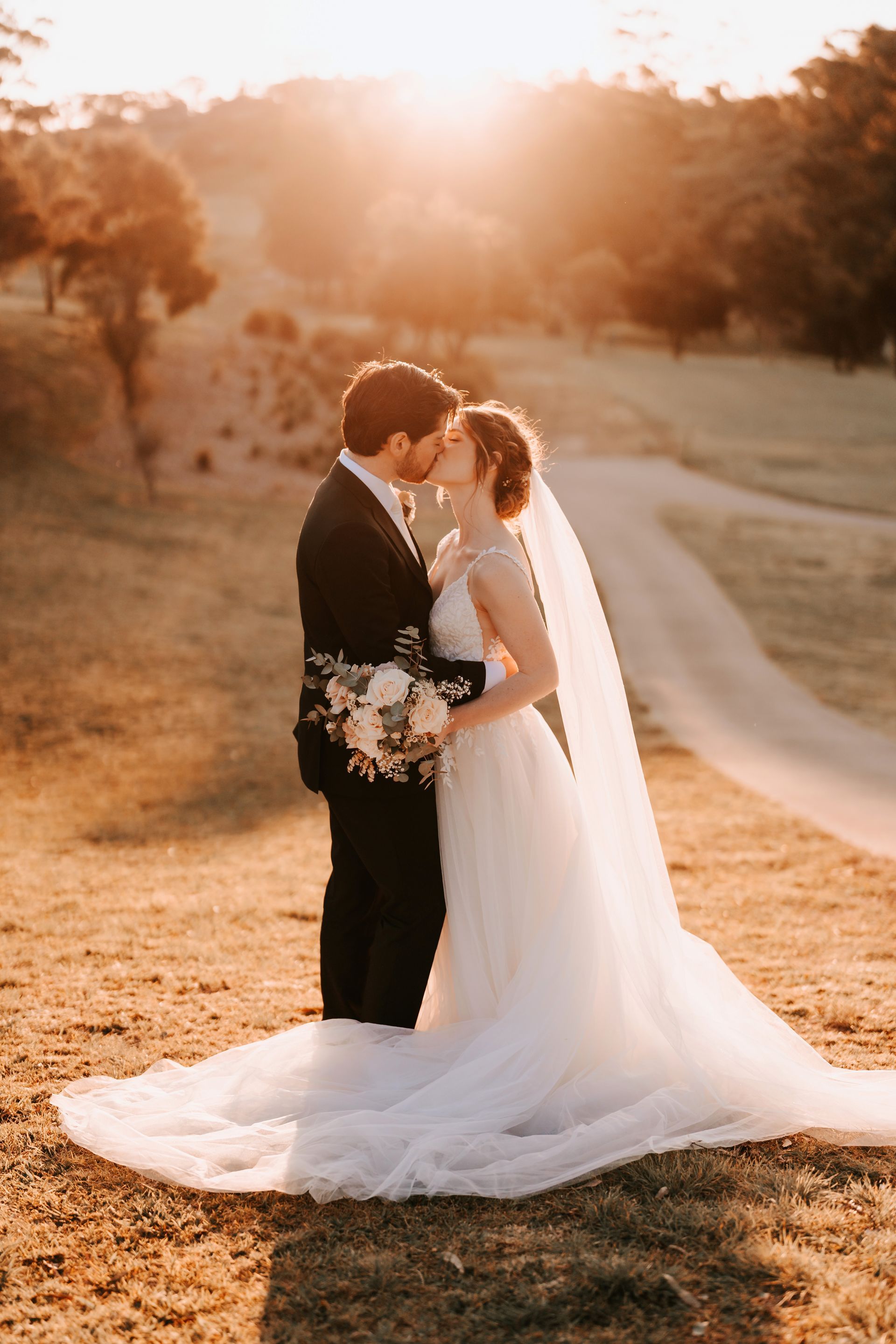 Couple kissing on a grassy hill at sunset, the bride in a white gown and veil, groom in a black suit.
