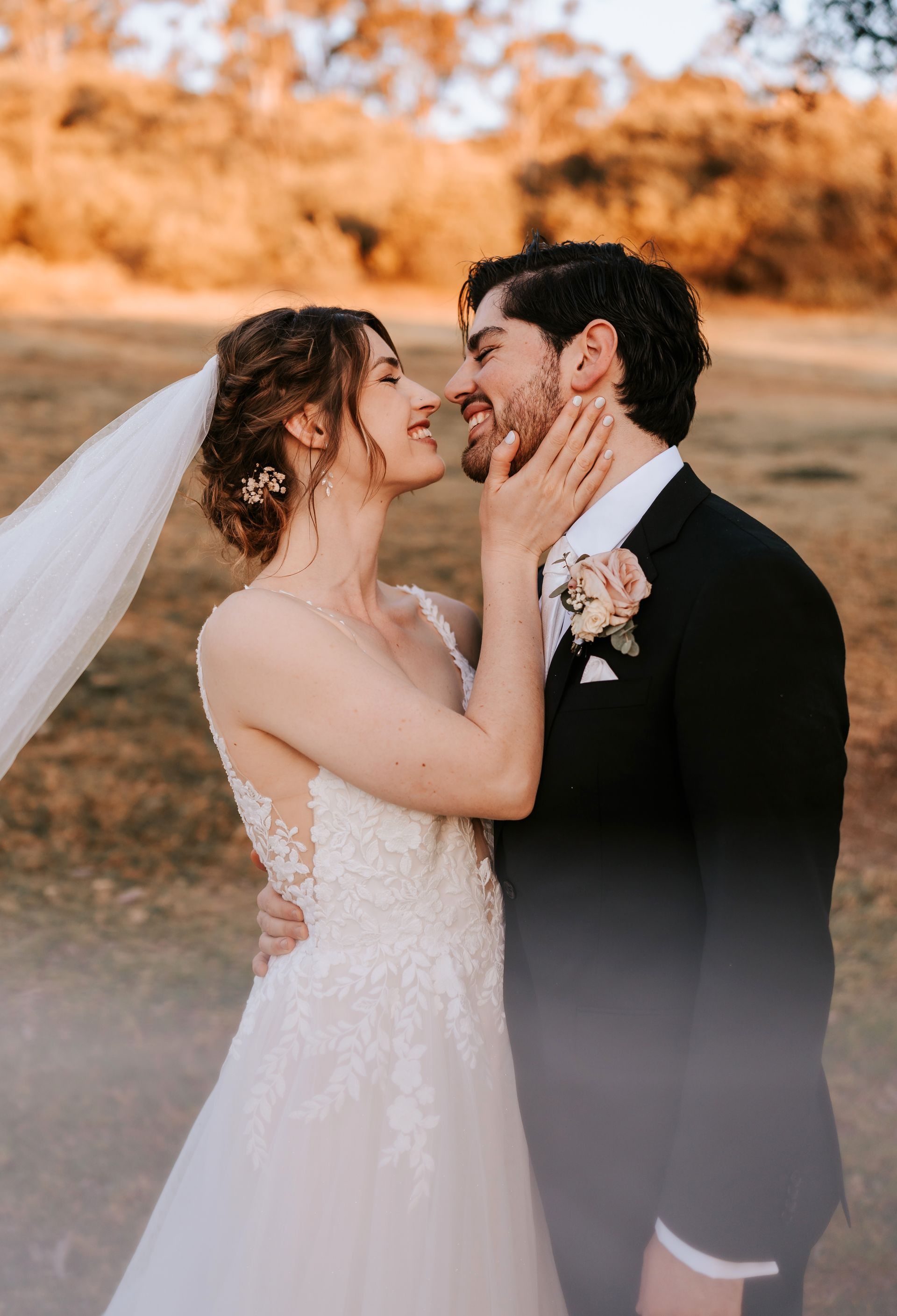 Bride and groom embrace outdoors; she touches his face, smiling. Soft sunlight, wedding attire.