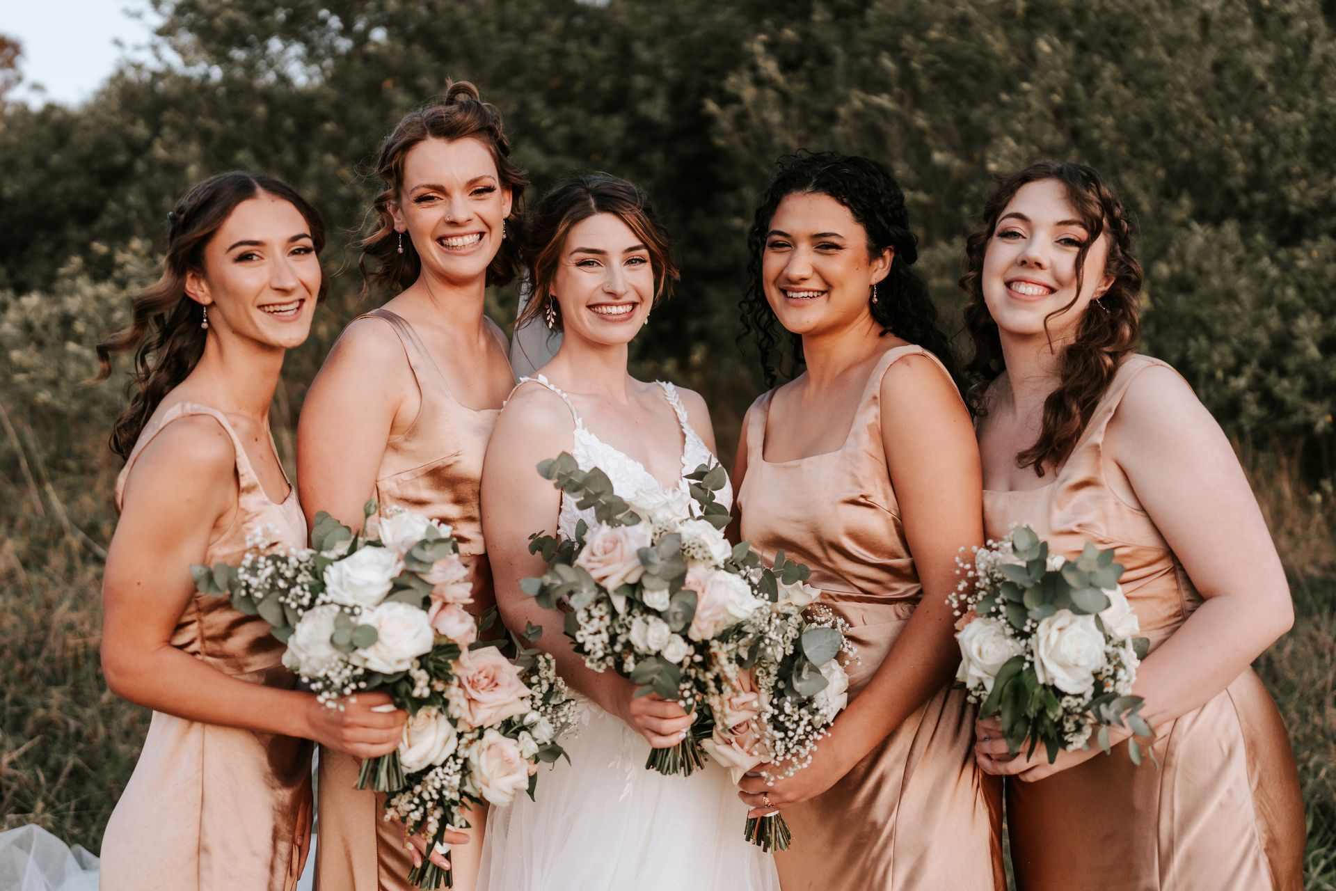 Bride and bridesmaids pose outdoors, holding bouquets. They wear peach-colored dresses and smile.