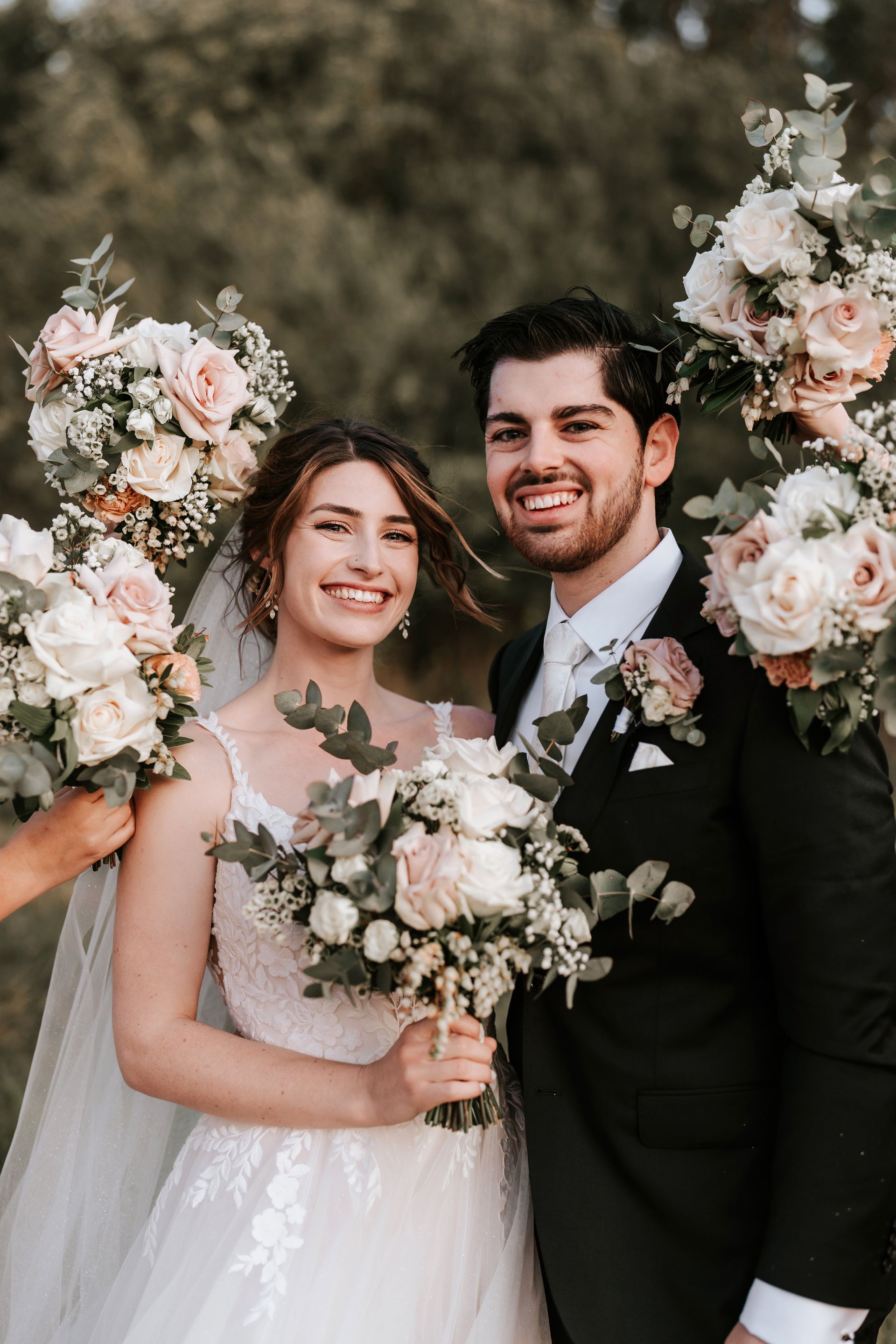 Bride and groom smiling at camera, surrounded by flower bouquets, outdoors.