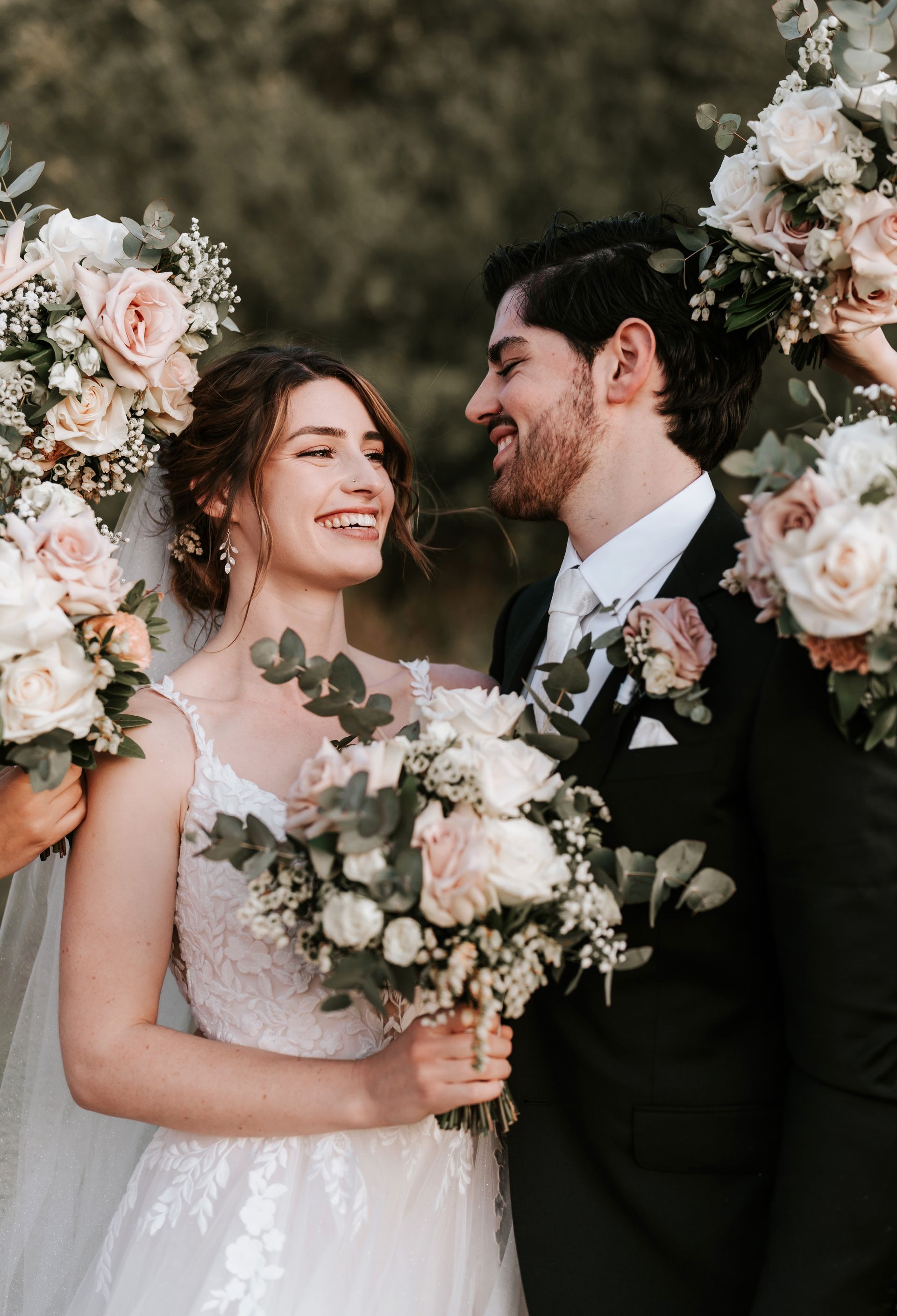 Bride and groom smiling, surrounded by bridesmaids with bouquets. Outdoor setting, soft lighting.