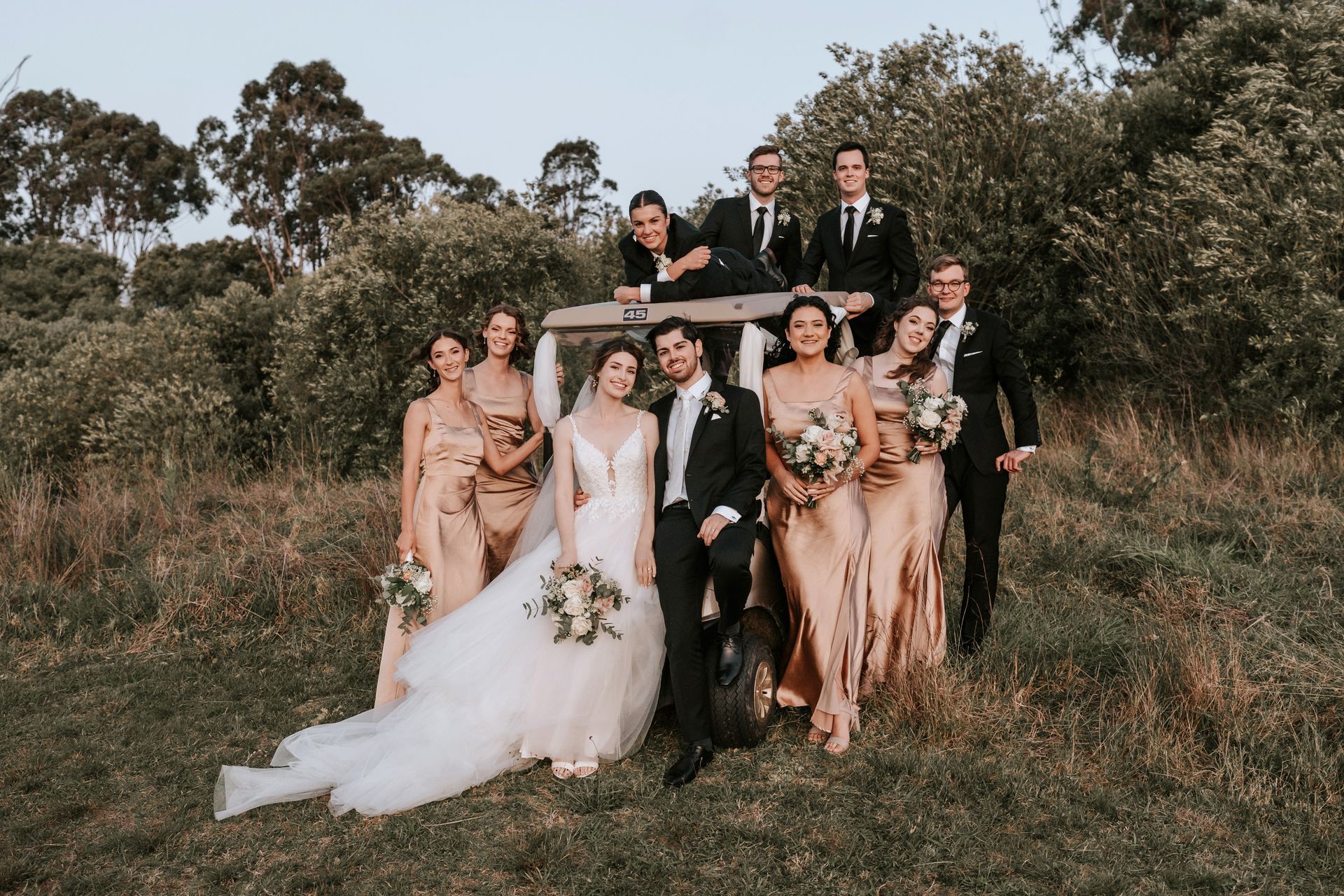 Wedding party poses near a vehicle in a field; bride and groom in front, bridesmaids in gold, groomsmen in black suits.