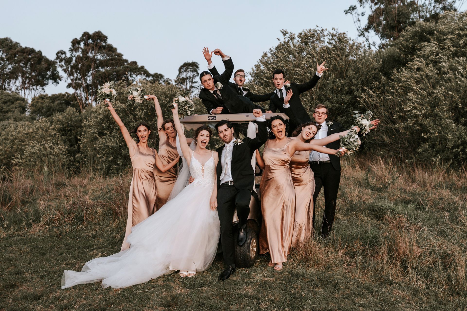 Wedding party celebrating outdoors. Bride and groom pose with bridesmaids and groomsmen, many with arms raised in joy.