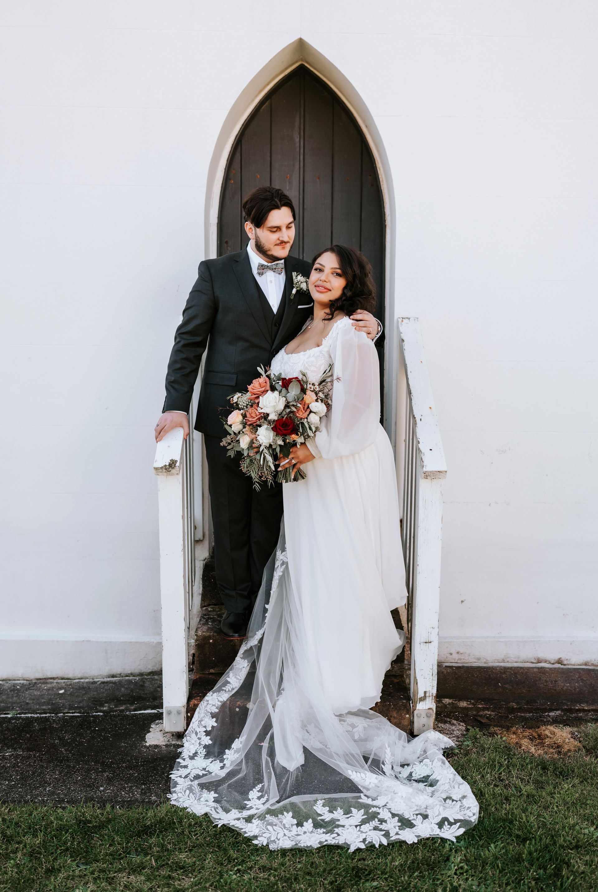 Wedding couple standing on steps of a church door; the bride holds a bouquet.