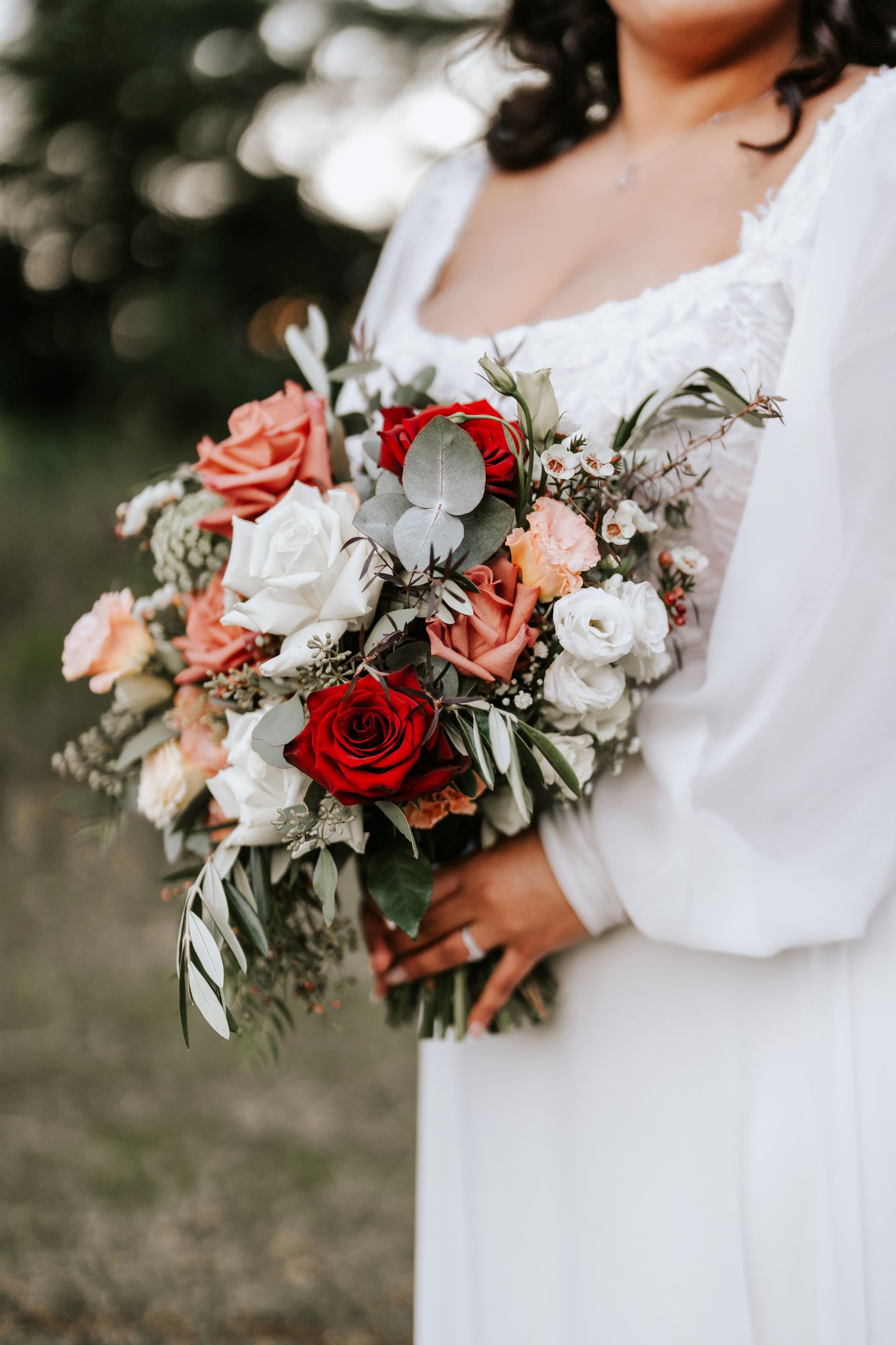Bride in white dress holding a bouquet of red, peach, and white flowers, outside.