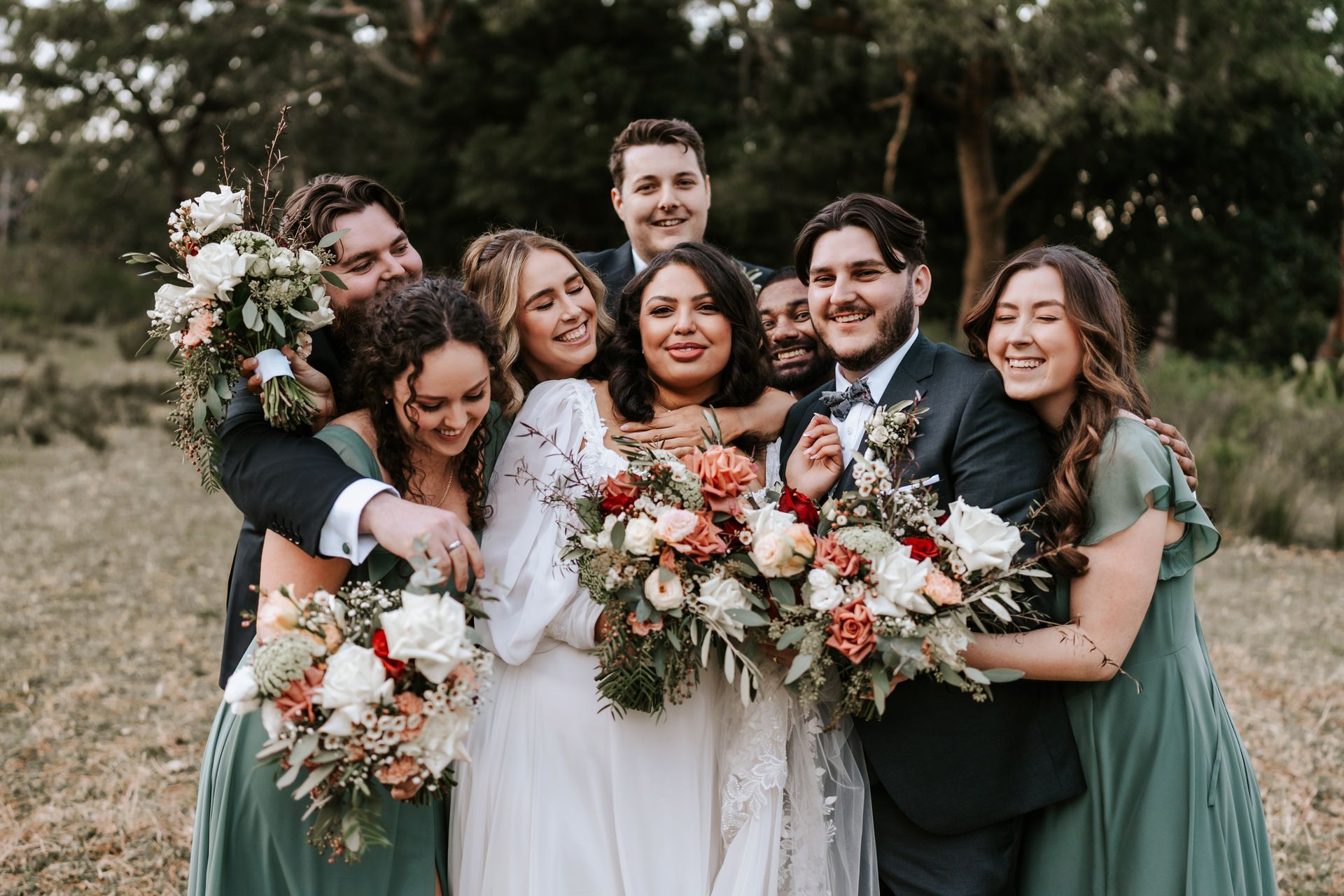 Wedding party: Bride surrounded by bridesmaids and groomsmen smiling in outdoor setting.