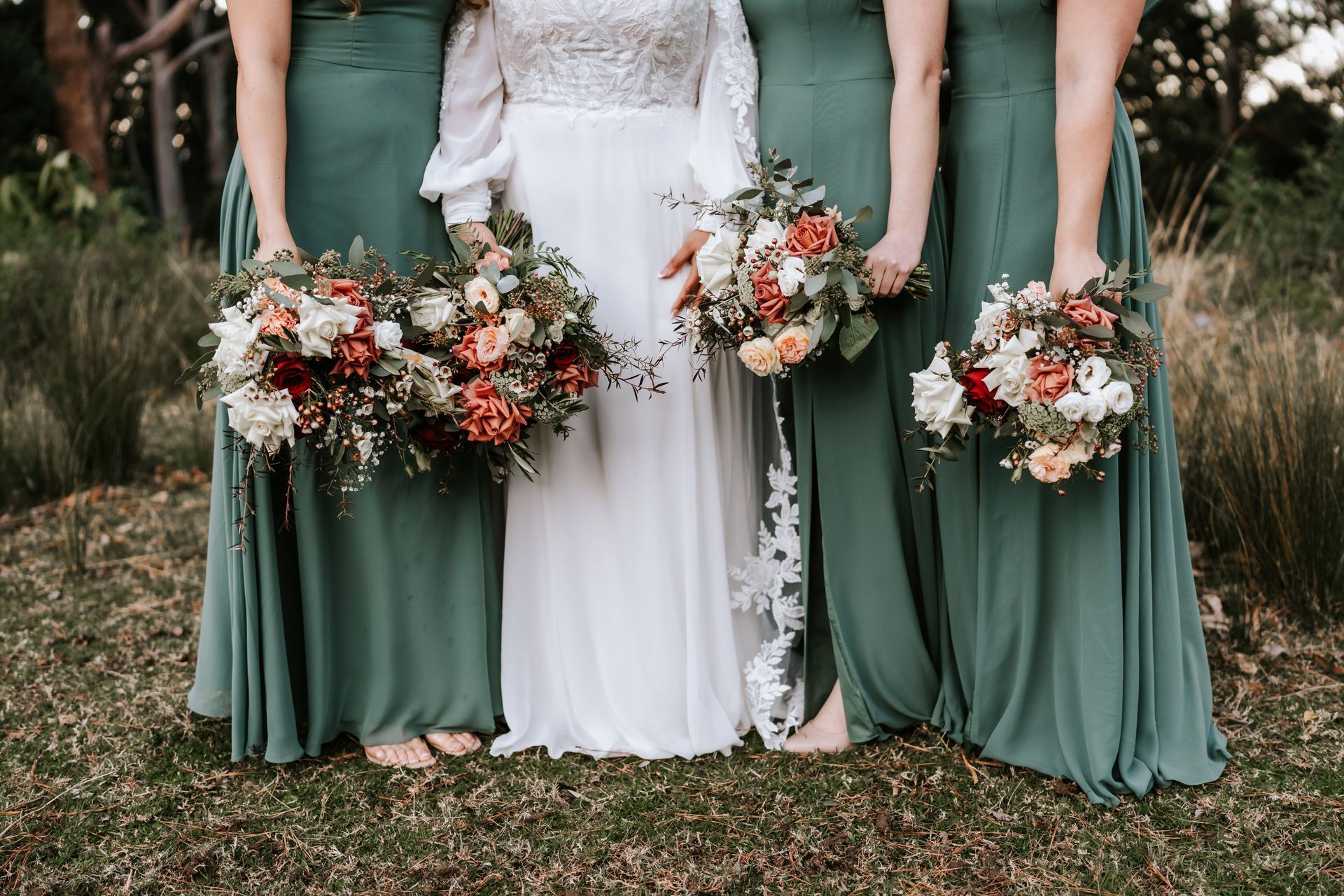 Bride in white gown, bridesmaids in green dresses hold bouquets on grass.