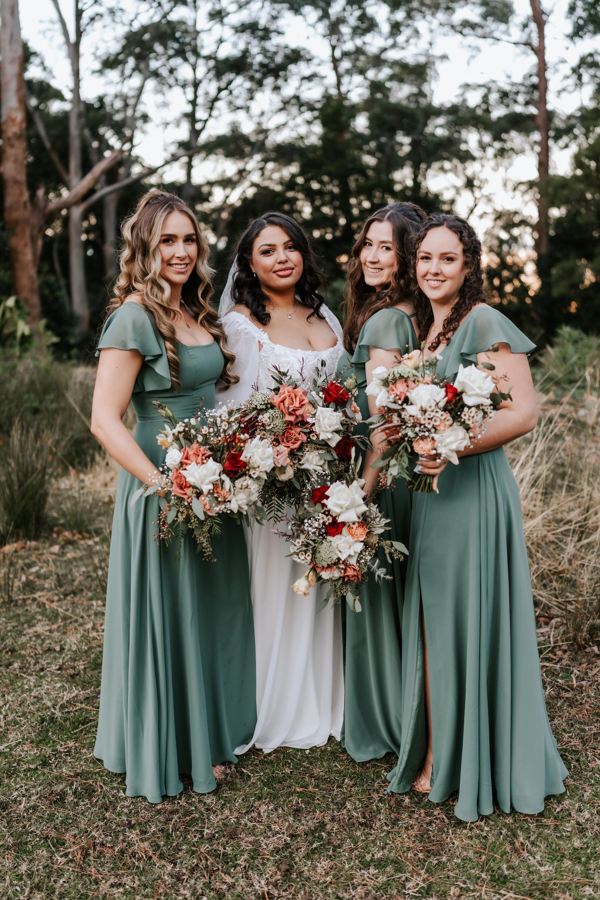 Bride with bridesmaids in a forest. Bride in white gown, bridesmaids in green dresses, holding bouquets.
