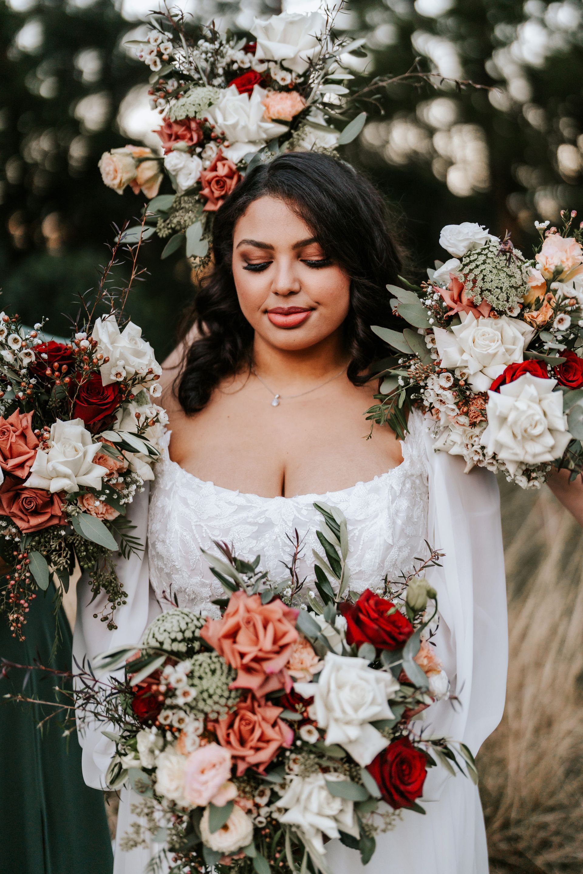 Bride in white dress, holding bouquets with red and white flowers, outdoors.