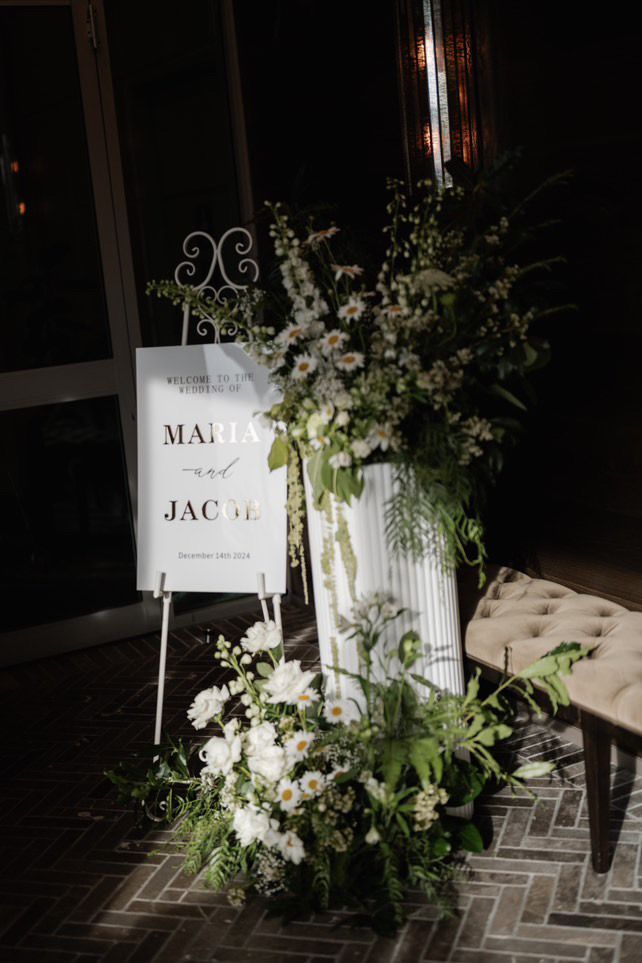 Wedding welcome sign with floral arrangements. White sign reads 
