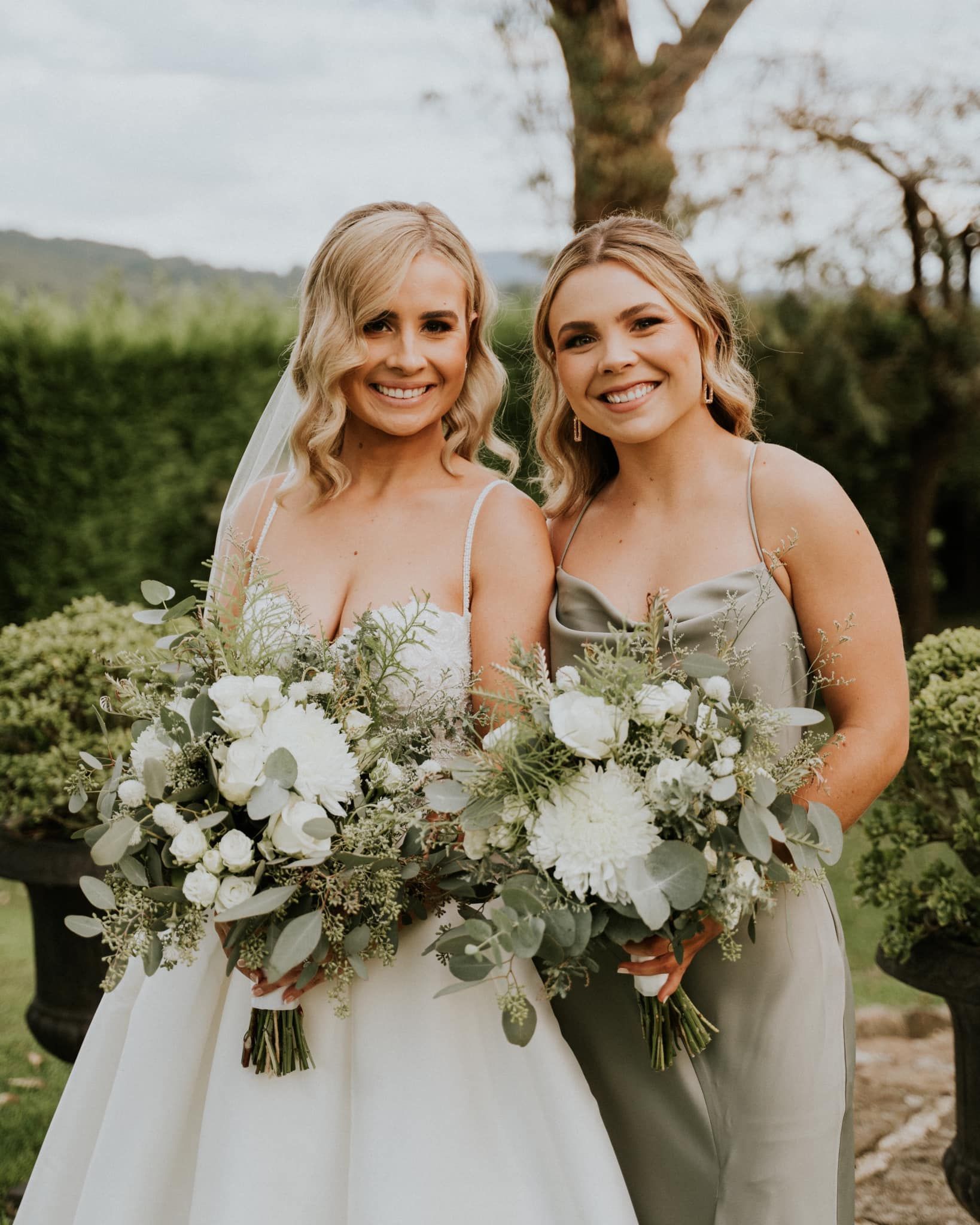 Bride and bridesmaid smiling, holding bouquets, outdoors. The bride wears a white dress, the bridesmaid wears green.