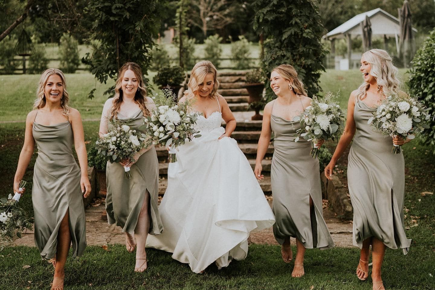 Bride in white gown with bridesmaids in green dresses, walking on grass.