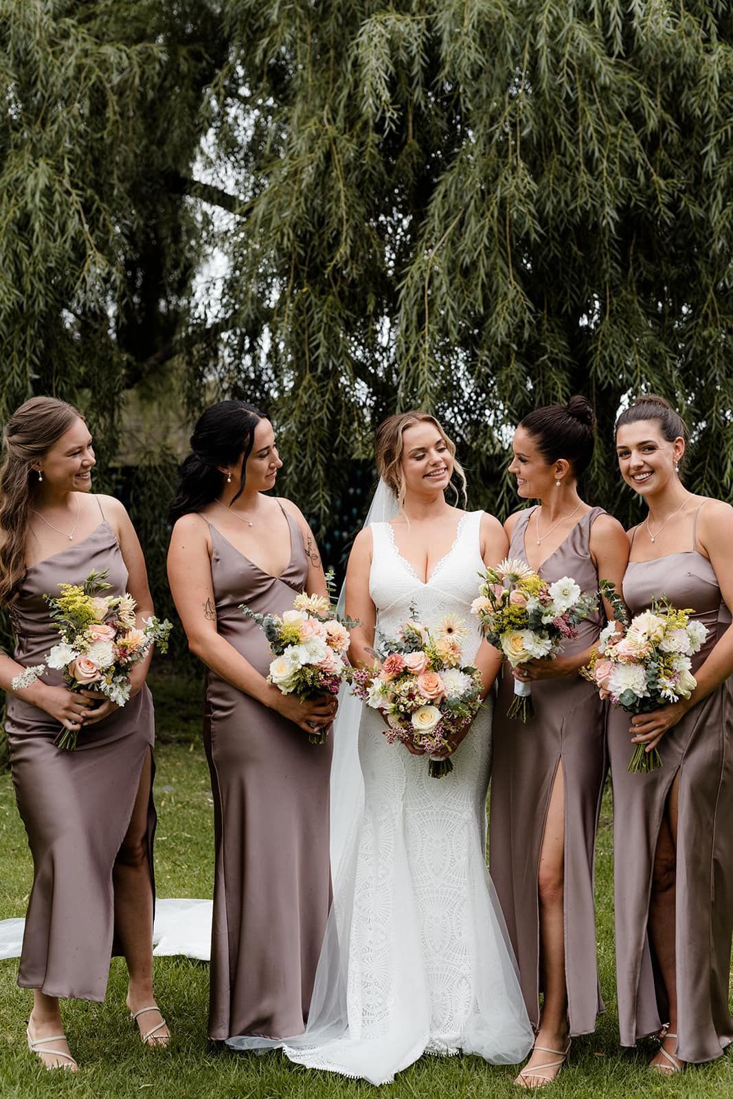 Bride and bridesmaids in dusty rose dresses holding bouquets, smiling in a garden.
