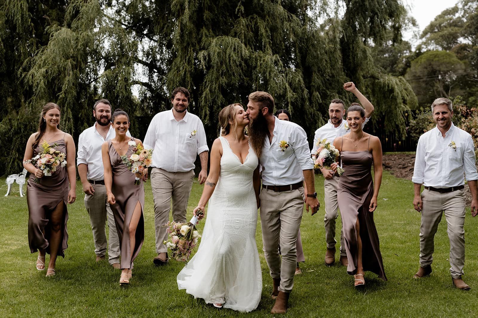 Wedding party outdoors. Bride and groom kiss. Others smile, some with arms raised. Green grass, trees.