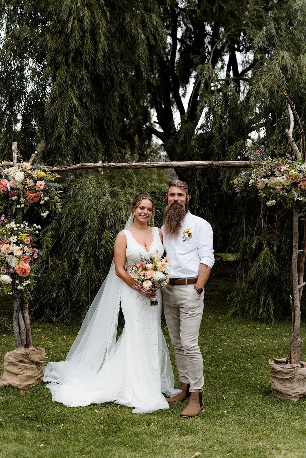 Bride and groom under a wooden arch, posing for a wedding photo outdoors.