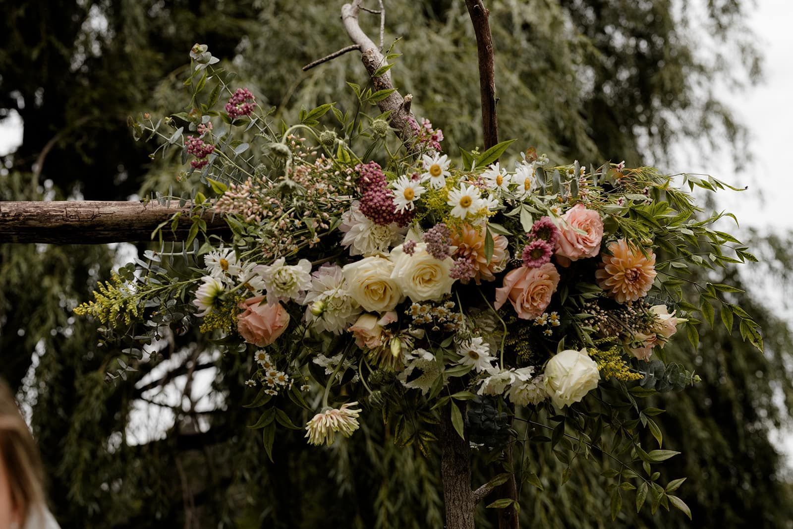 Floral arrangement on a wooden arch, with pink, white, and yellow flowers against a green backdrop.