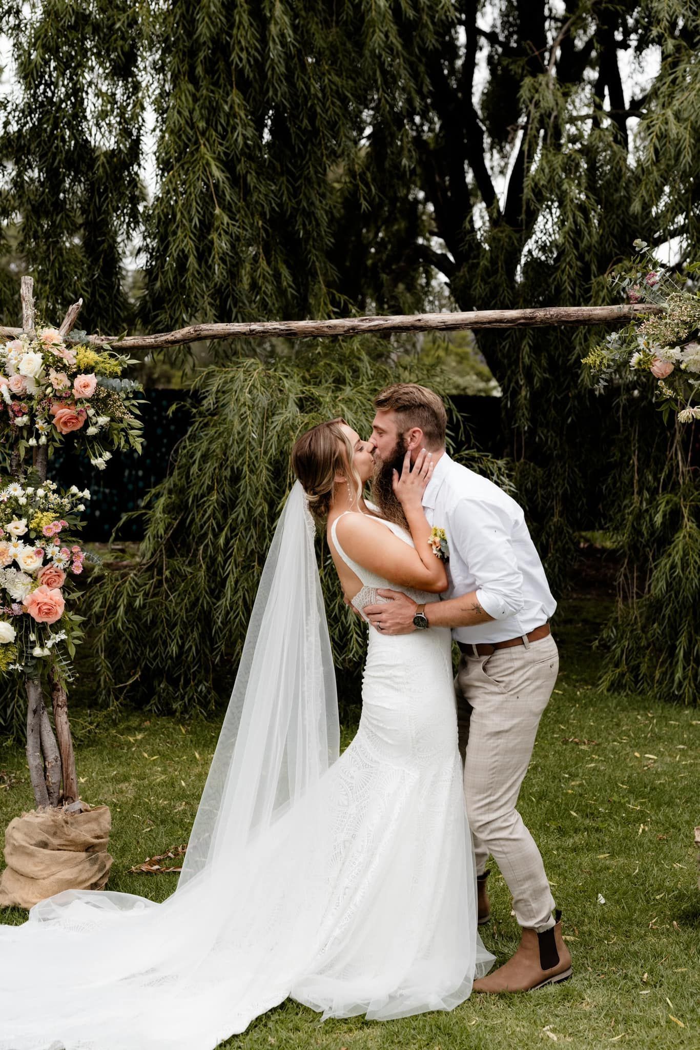 Bride and groom kissing under a floral archway during a wedding ceremony in a garden.