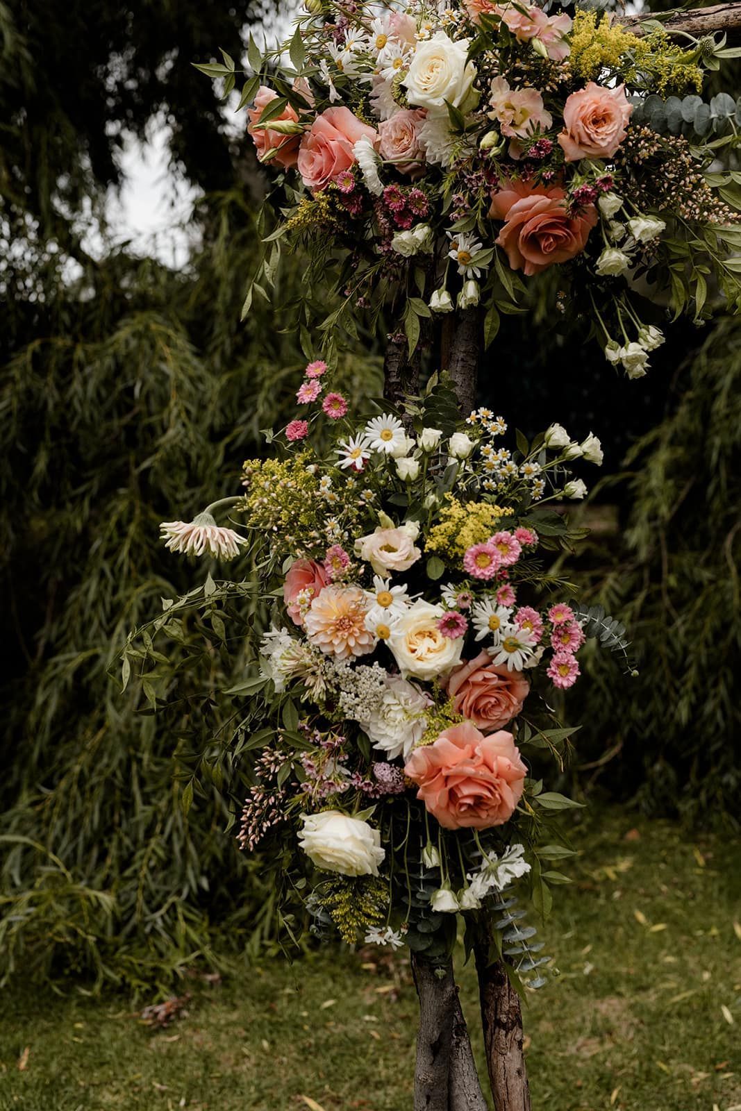 Floral arrangement on a tree trunk with peach roses, white flowers, and green foliage in an outdoor setting.