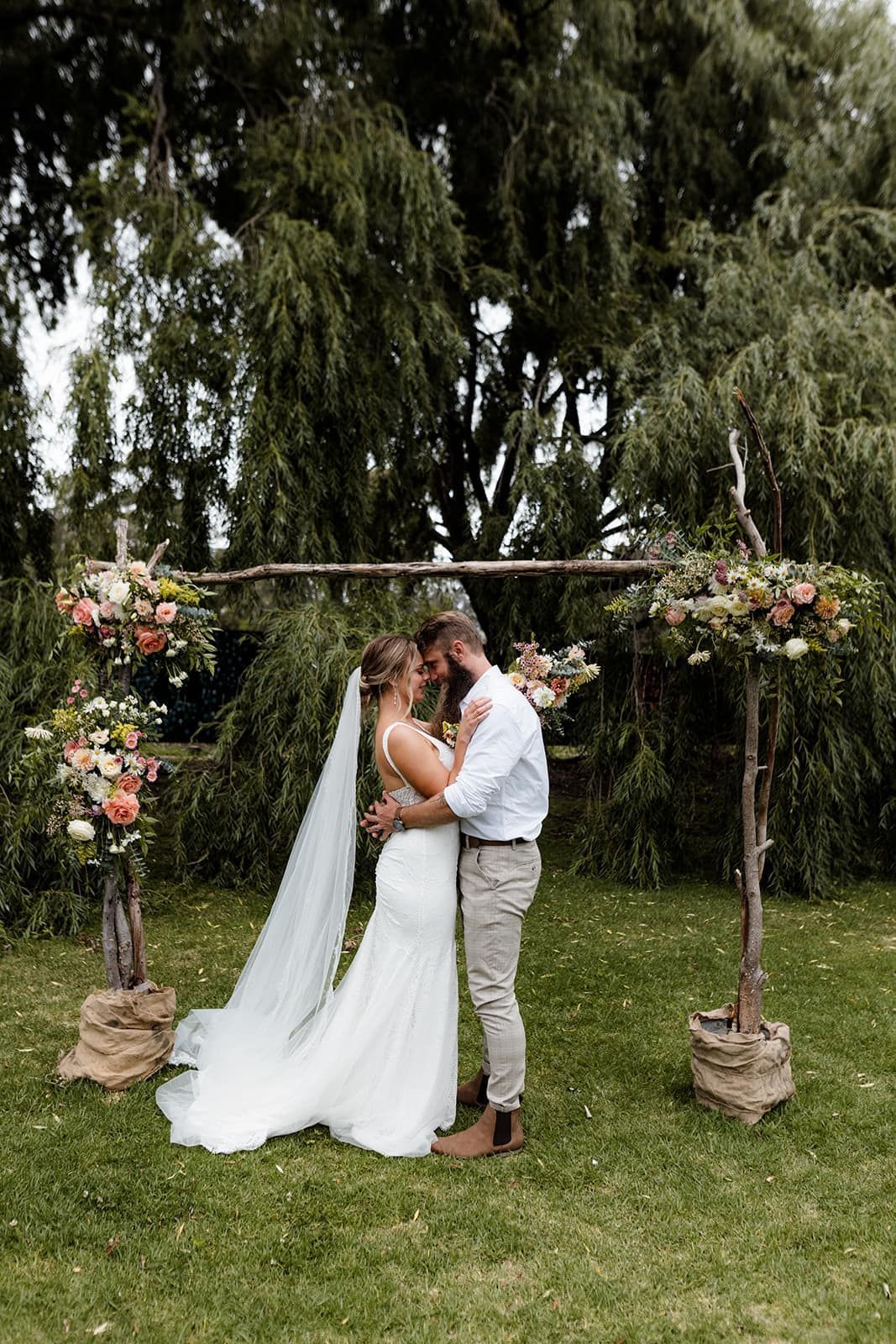 Couple embraces under a flower-decorated archway during an outdoor wedding ceremony.