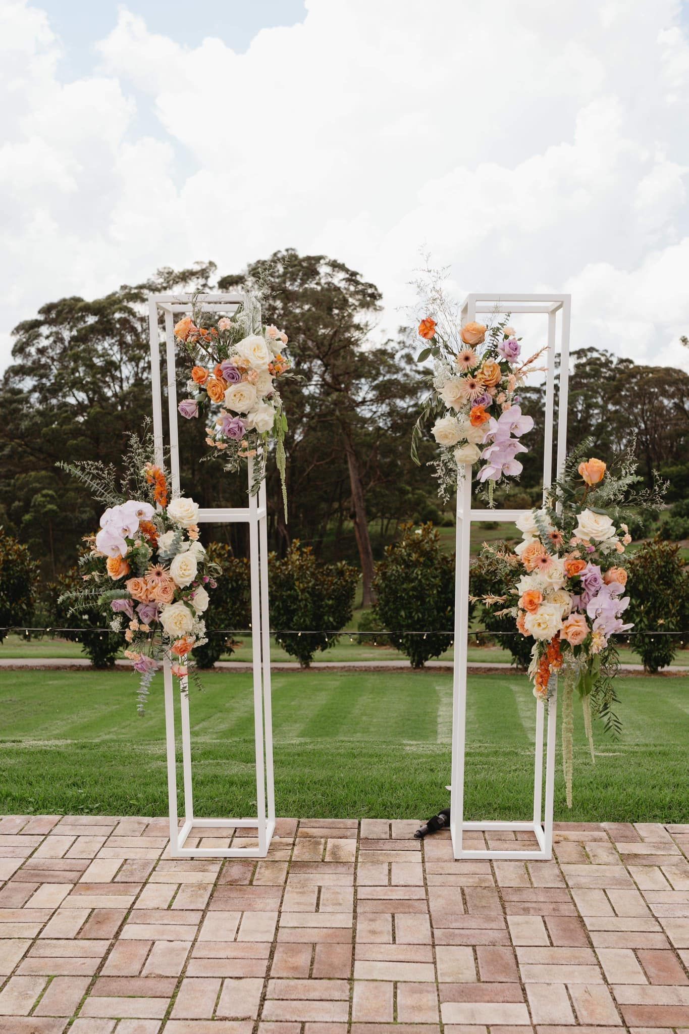 White geometric wedding arch with colorful floral arrangements outdoors on brick and grass.
