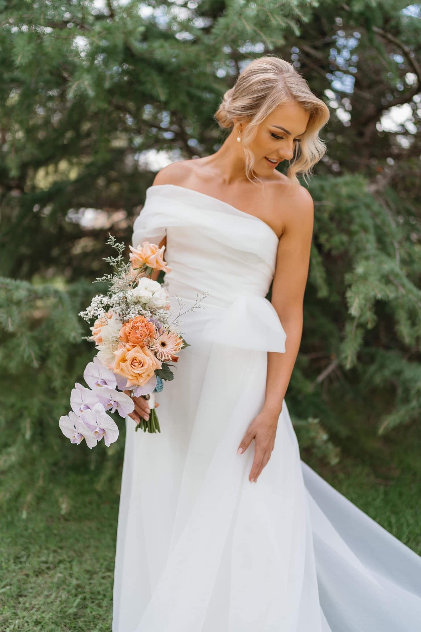Blonde bride in white off-shoulder dress, holding flowers, smiling outdoors.
