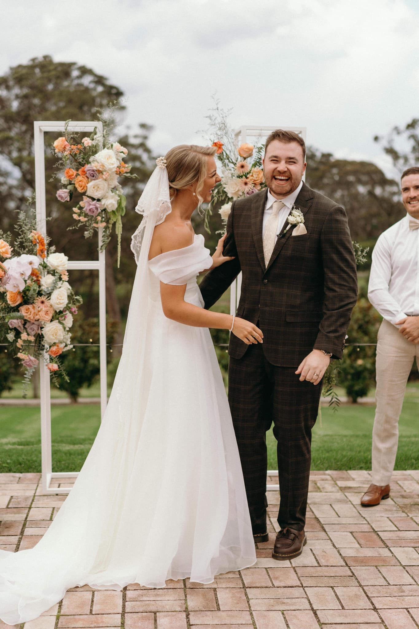 Bride and groom smile at each other at wedding ceremony, standing before floral arch.