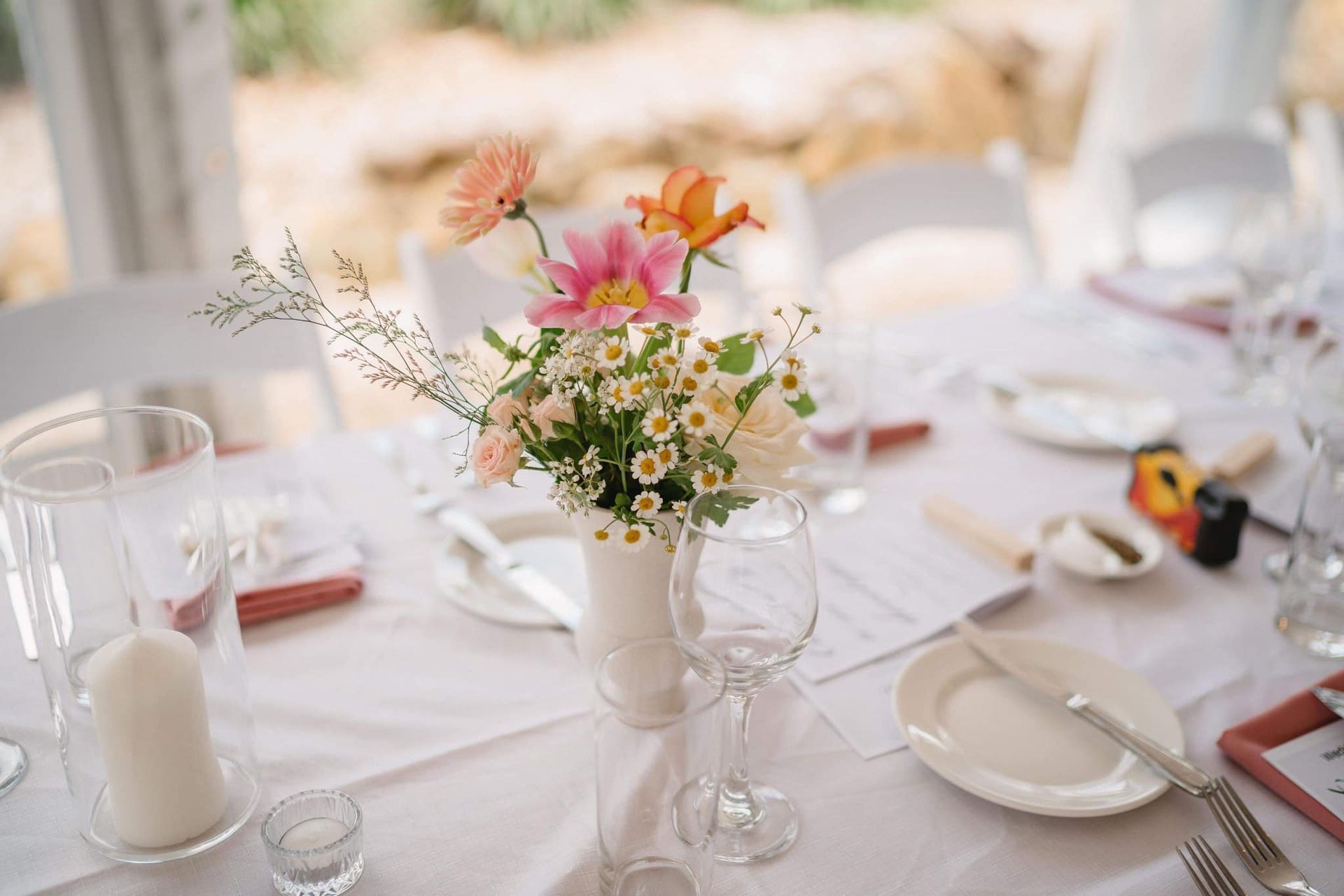 Floral centerpiece on a white tablecloth, set for a meal with white chairs and a blurred outdoor background.