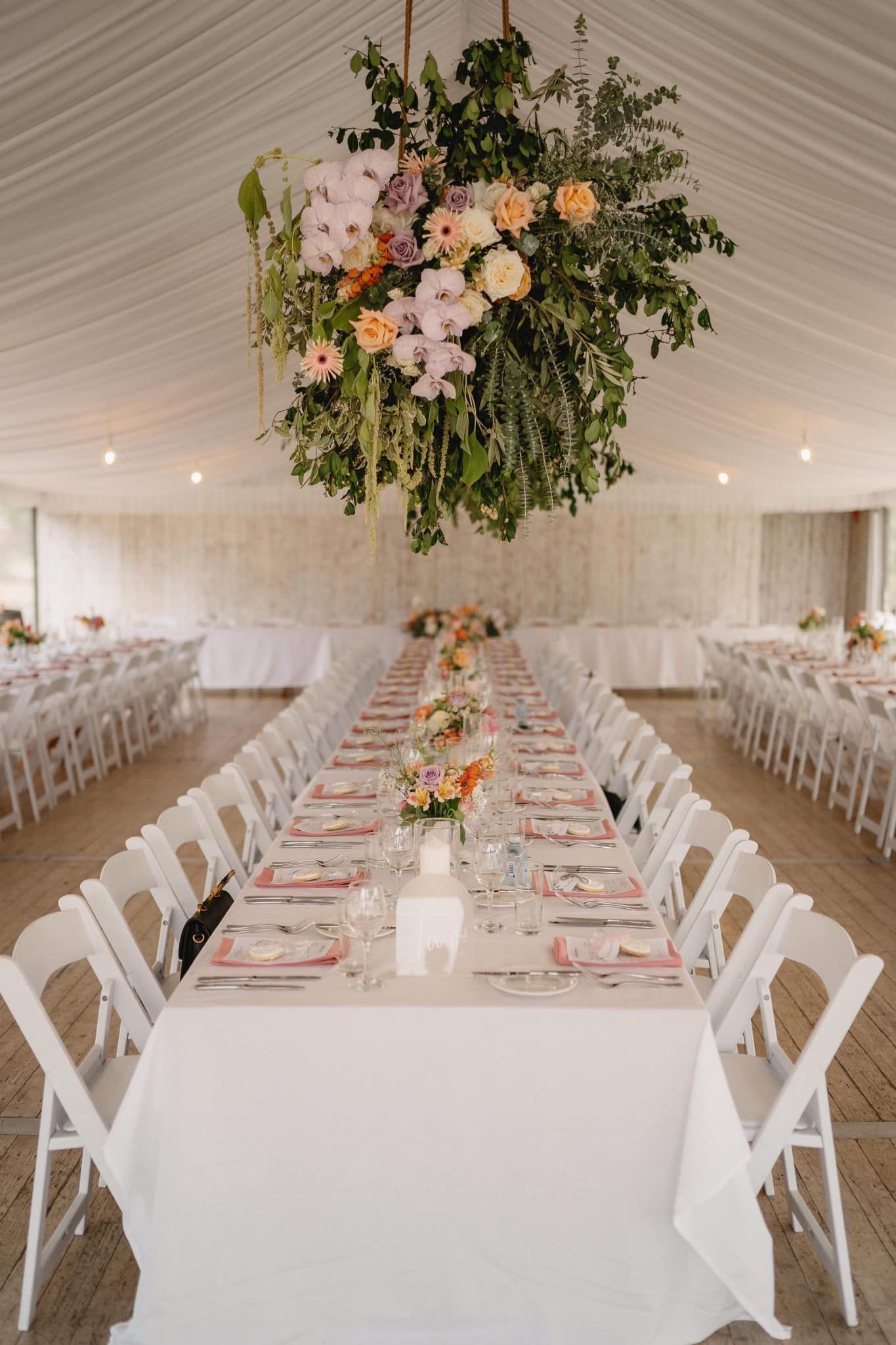 Long white table set for a wedding reception, with flowers and hanging floral arrangement.