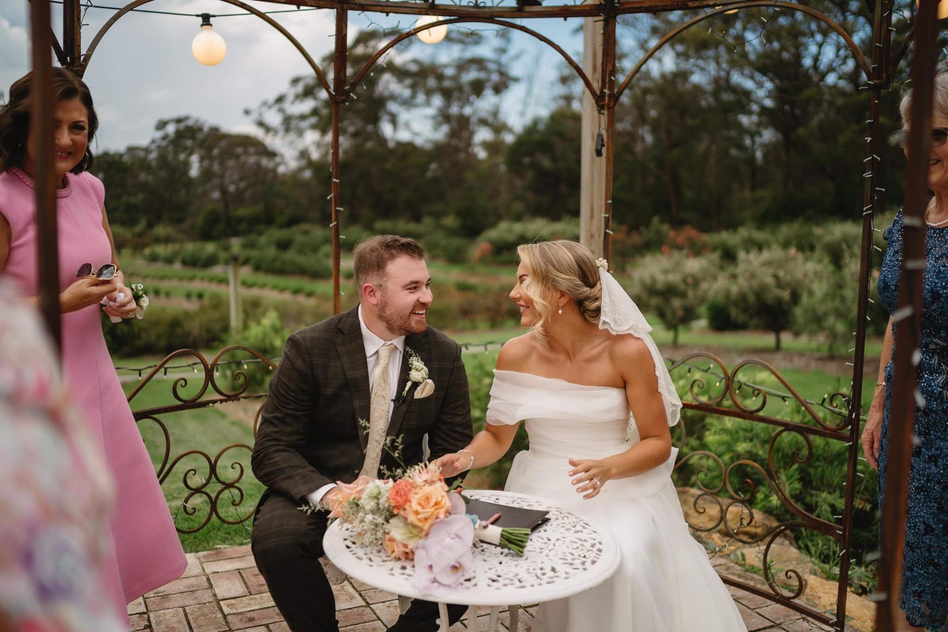 Bride and groom seated at a table in an outdoor wedding ceremony, smiling at each other.