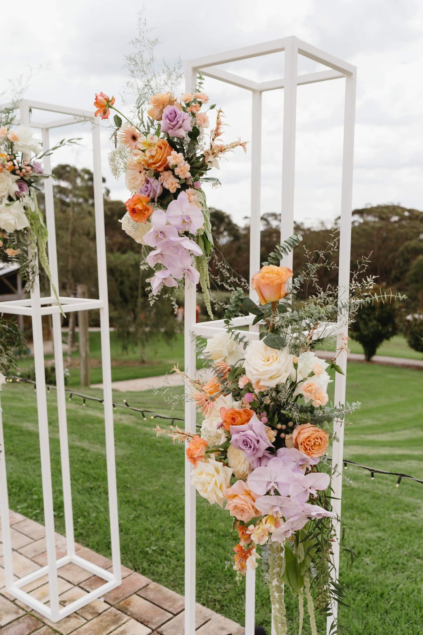 White floral arch with colorful flowers set on a green lawn with trees in the background.