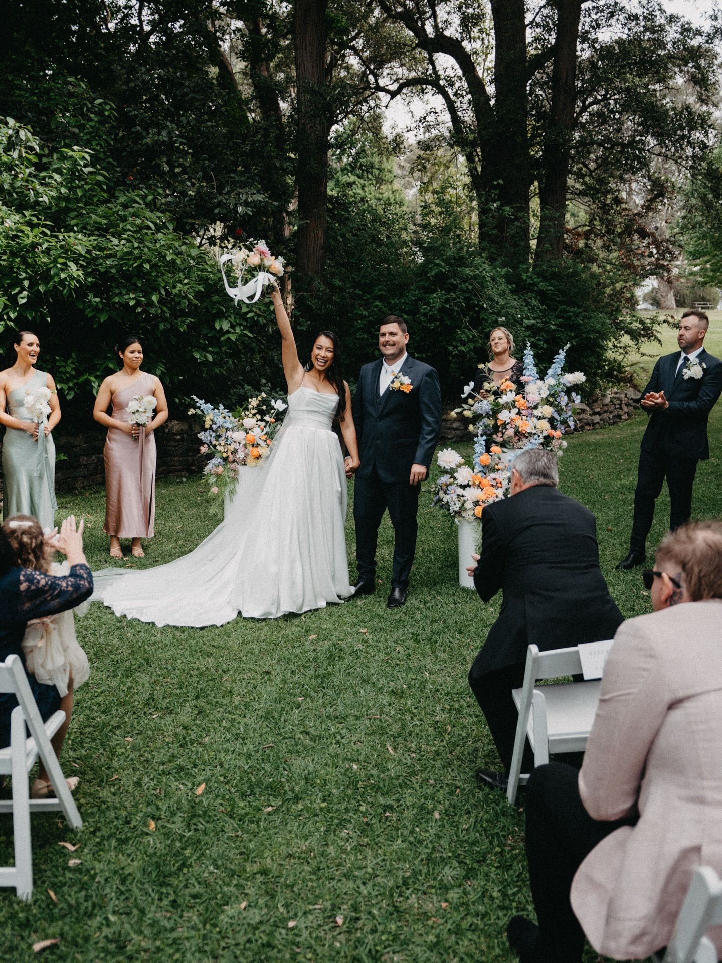 Newlyweds celebrate, bride raising bouquet, groom smiling, surrounded by wedding party and guests in garden.