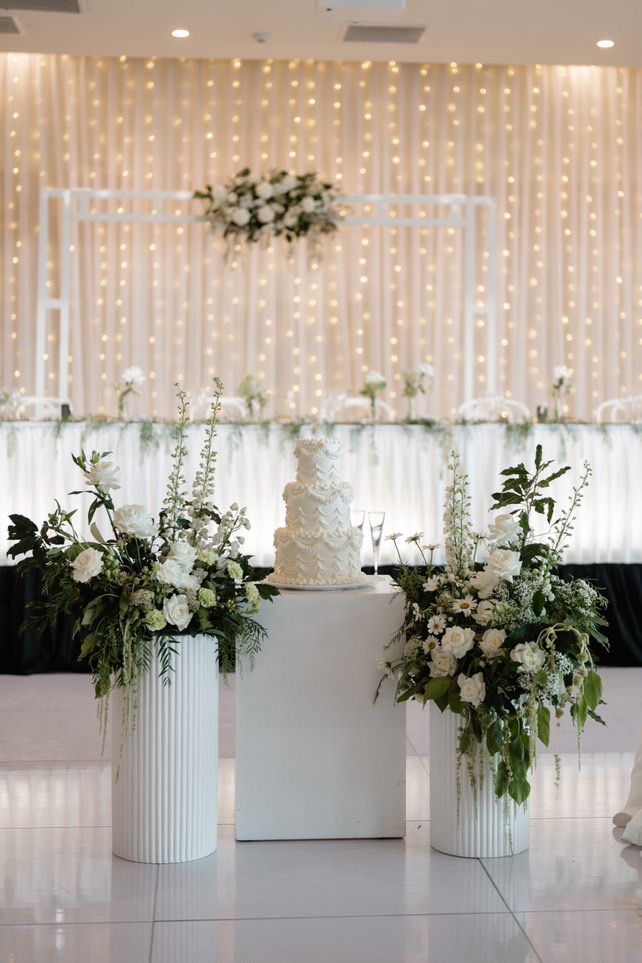Wedding cake on white pedestal with floral arrangements, in front of decorated backdrop.