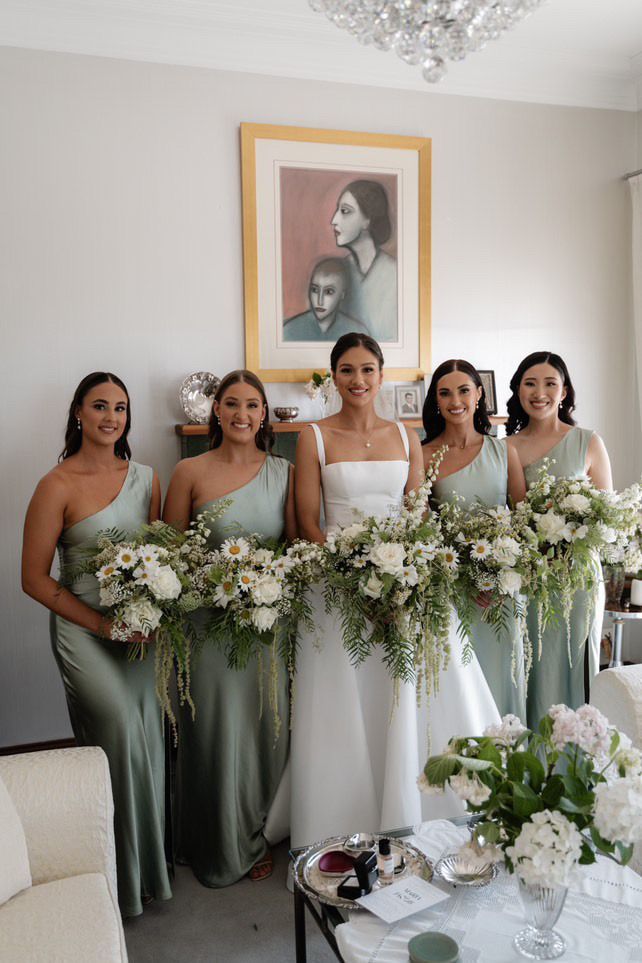 Bride and bridesmaids in sage green gowns holding bouquets. Bride in white dress, smiling. Indoor setting.