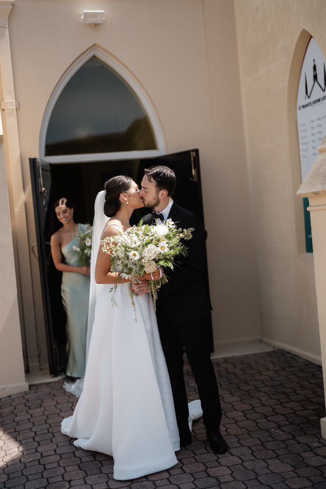 Bride and groom kissing outside a church, holding flowers. Bridesmaid in the background.