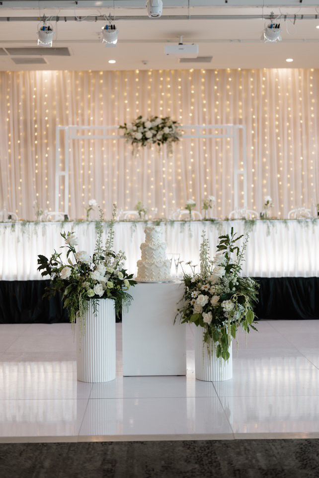 Wedding reception setup with a cake on a white table, floral arrangements, and a decorated backdrop with lights.