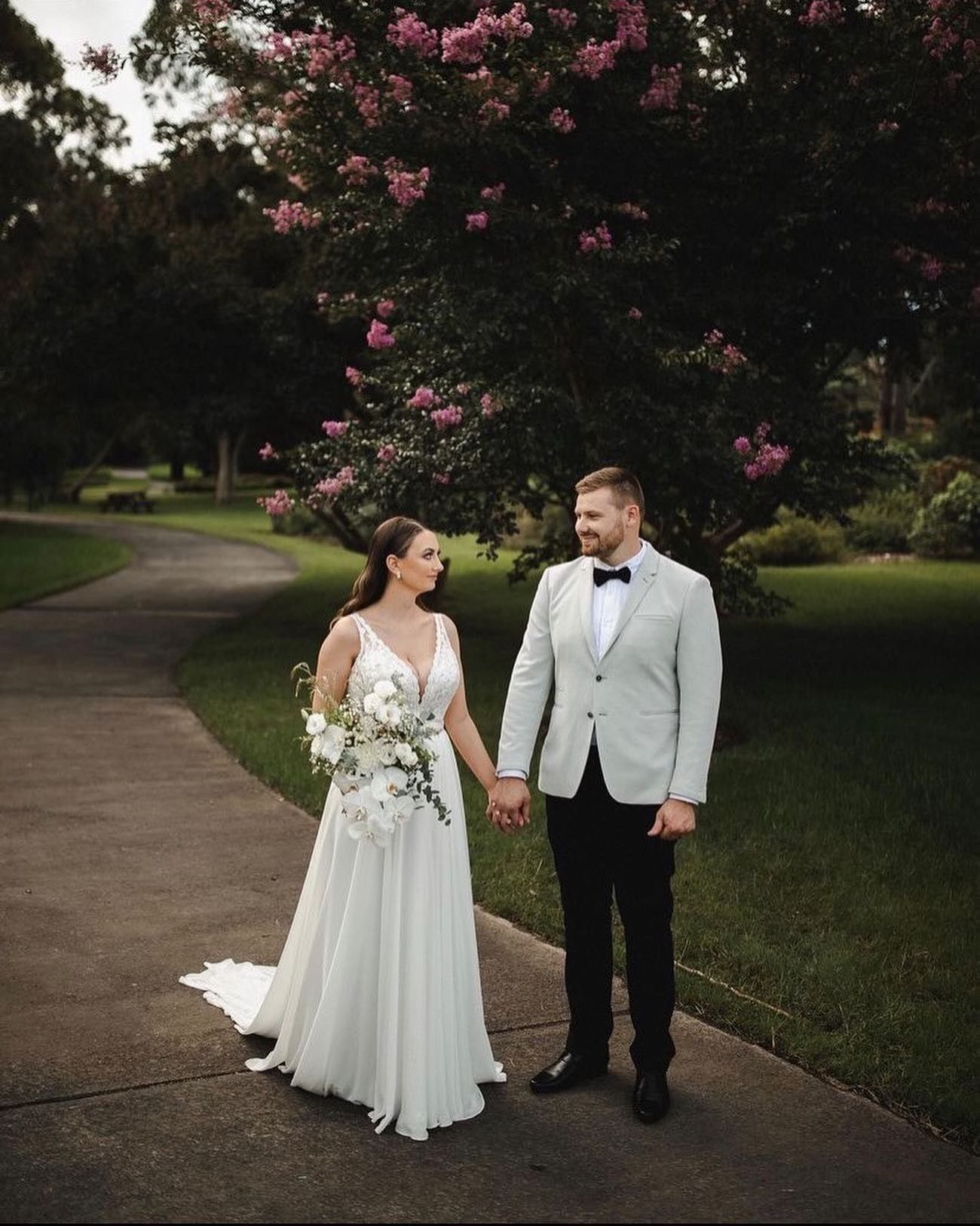 Bride and groom holding hands on a paved path under a flowering tree, looking at each other.