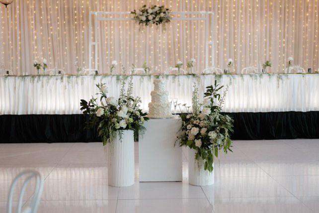 Wedding reception: cake on a white stand, floral arrangements, backdrop with lights, white and black decor.