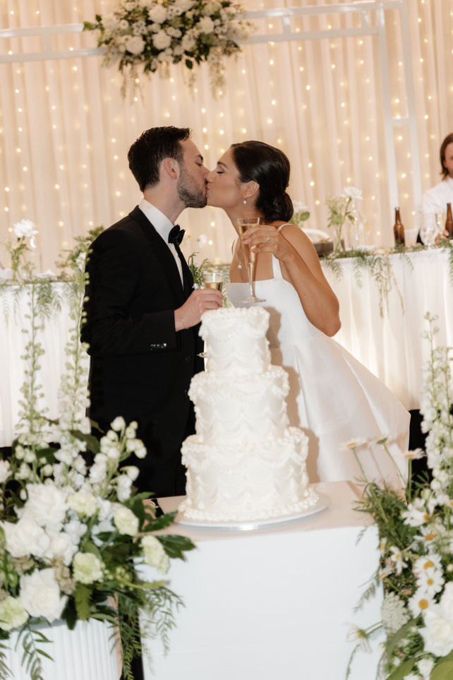 Couple kissing over a white wedding cake at a reception, surrounded by flowers and lights.