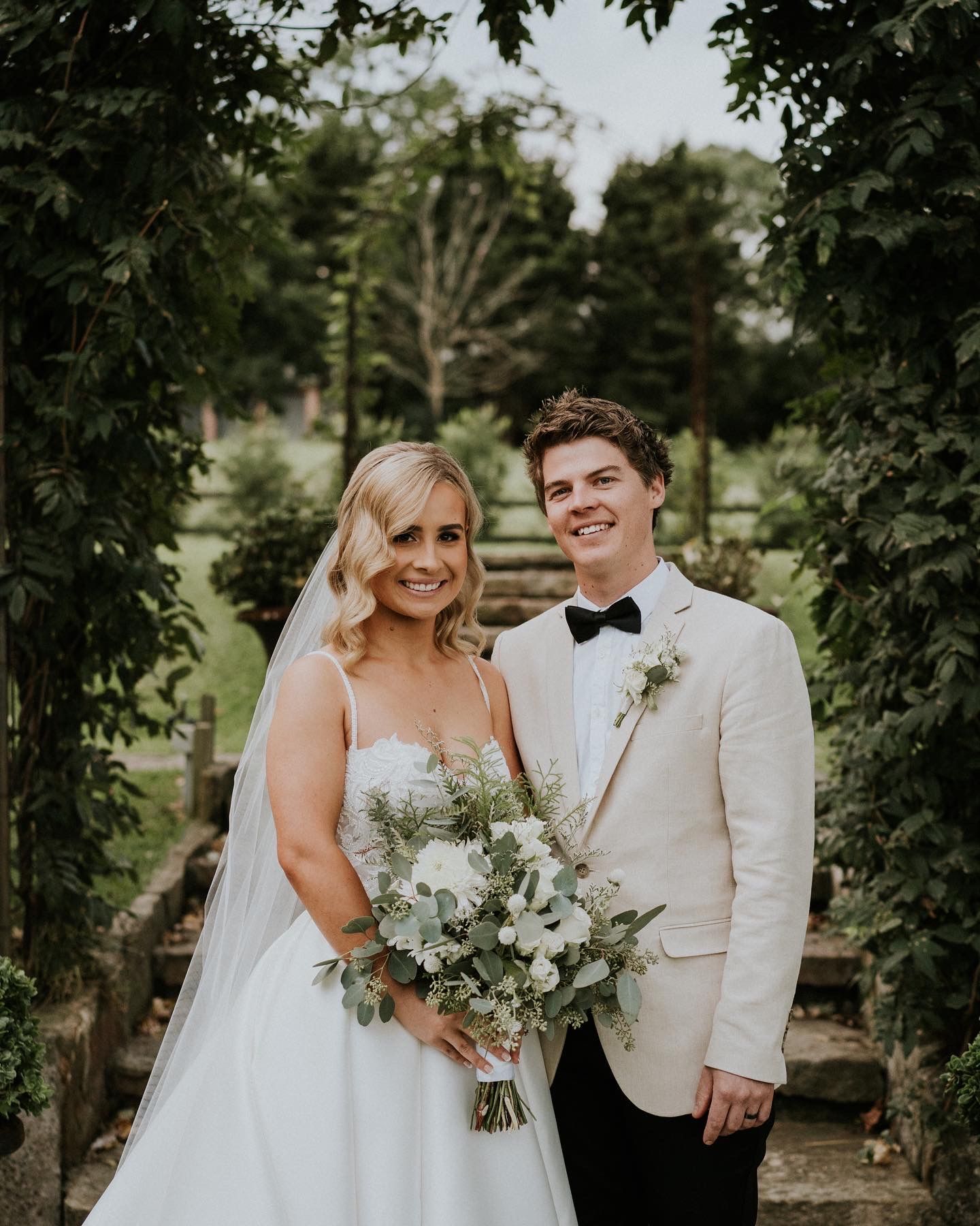 Bride and groom pose on steps under a floral archway, smiling. The bride holds a bouquet, the groom wears a bow tie.