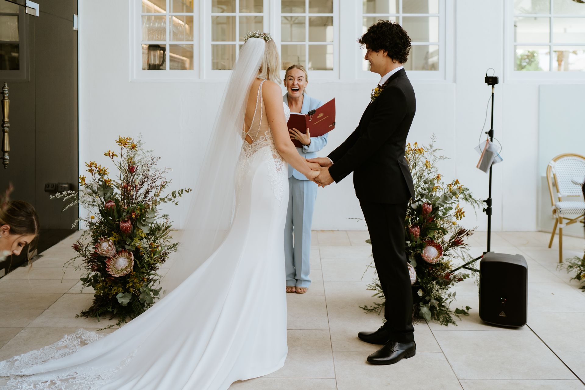 Couple holding hands during wedding ceremony, officiant smiles, flowers and speaker on white background.