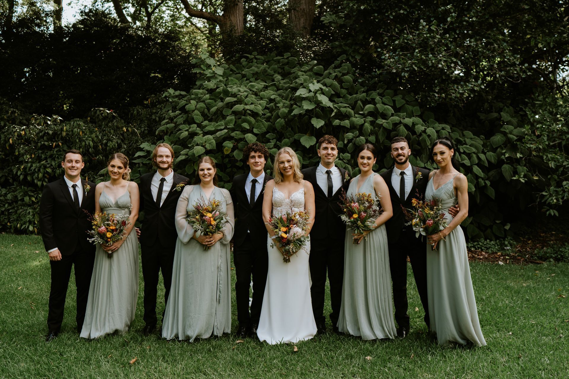 Wedding party, a bride and groom with bridesmaids and groomsmen in a green outdoor setting.