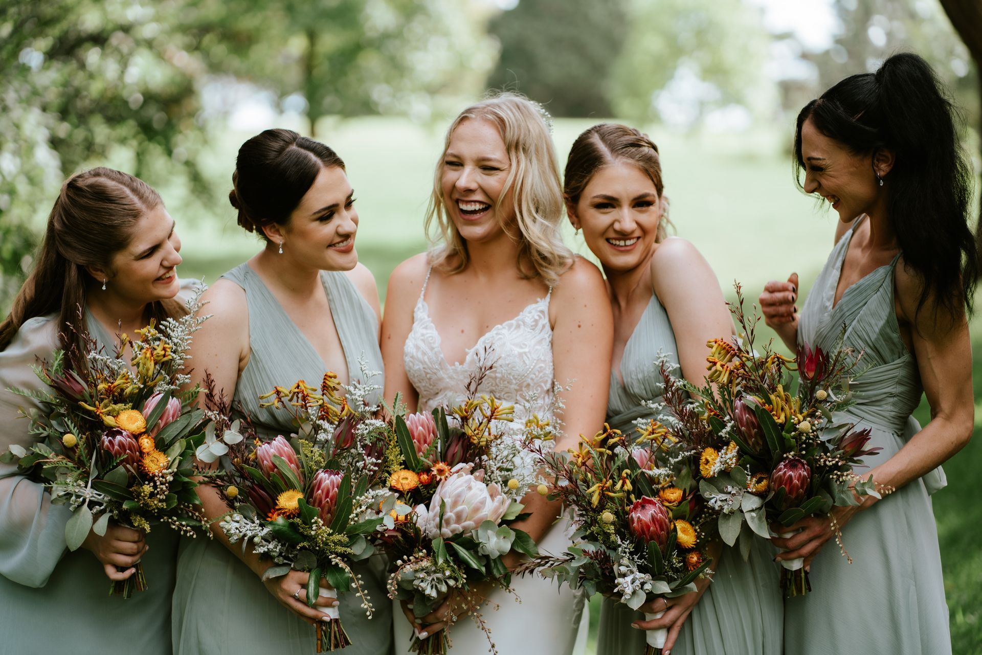 Bride and bridesmaids laughing outdoors, holding bouquets. They wear dresses in shades of sage green.