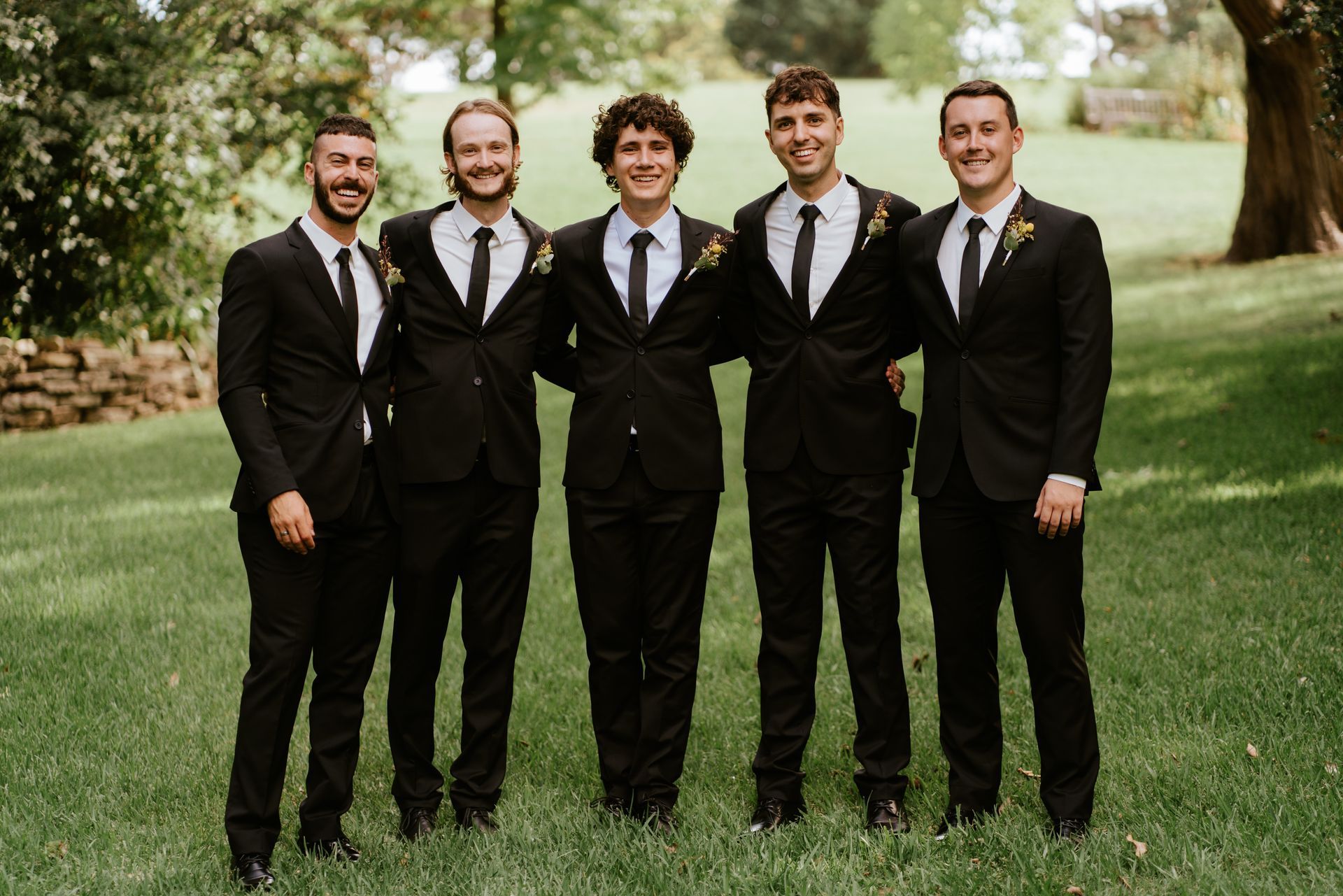 Five men in black suits and ties pose on grass, arms around each other, smiling.